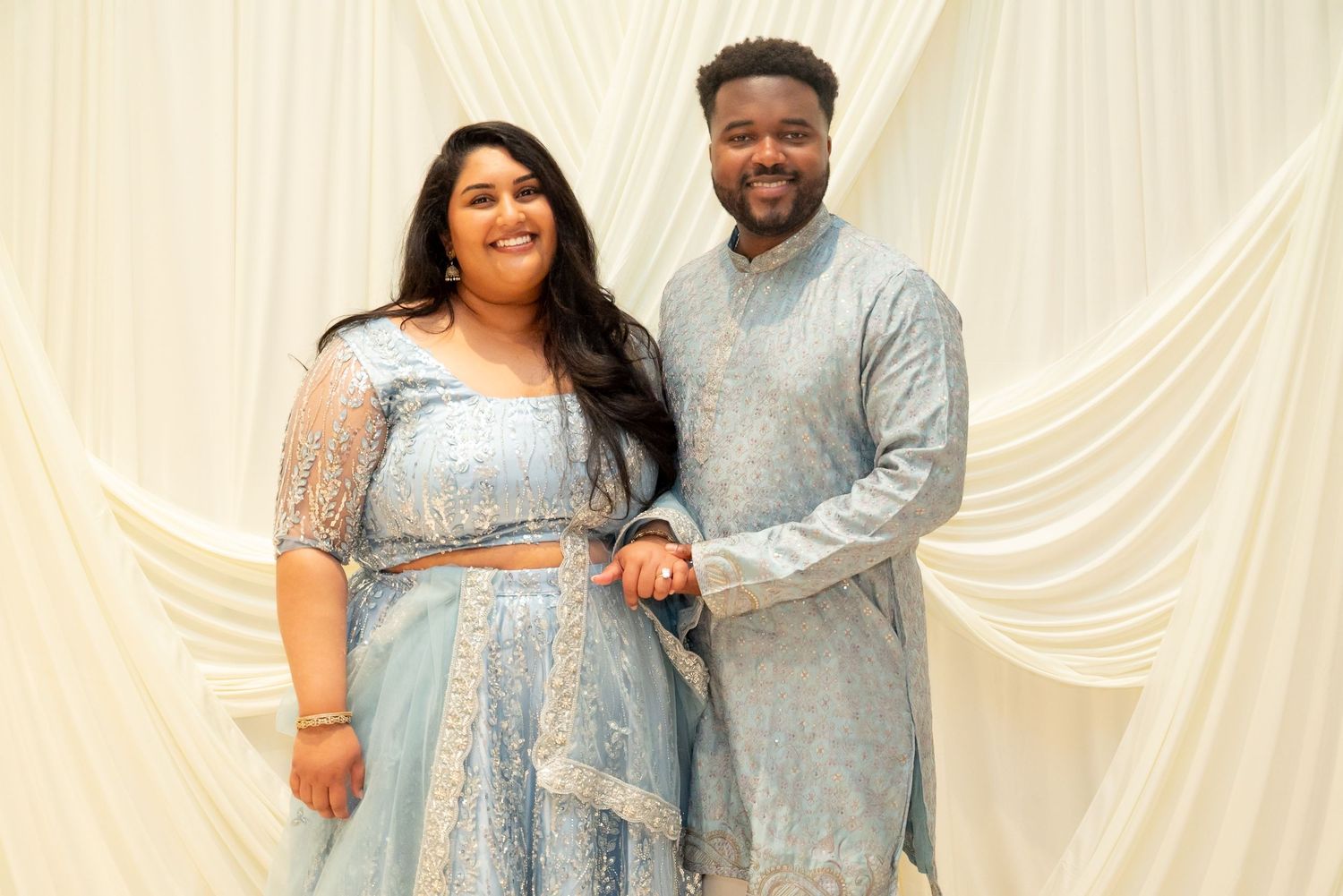 Couple in matching light blue sequined Indian formal wear posing together against white draped backdrop.