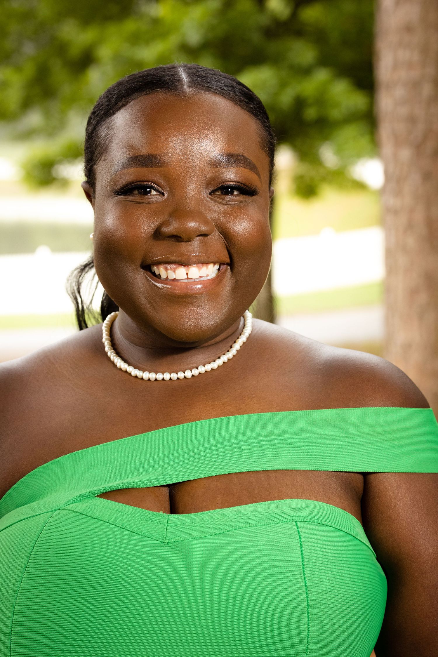 Cheerful woman in bright green off-shoulder top and pearl necklace smiling outdoors near trees.