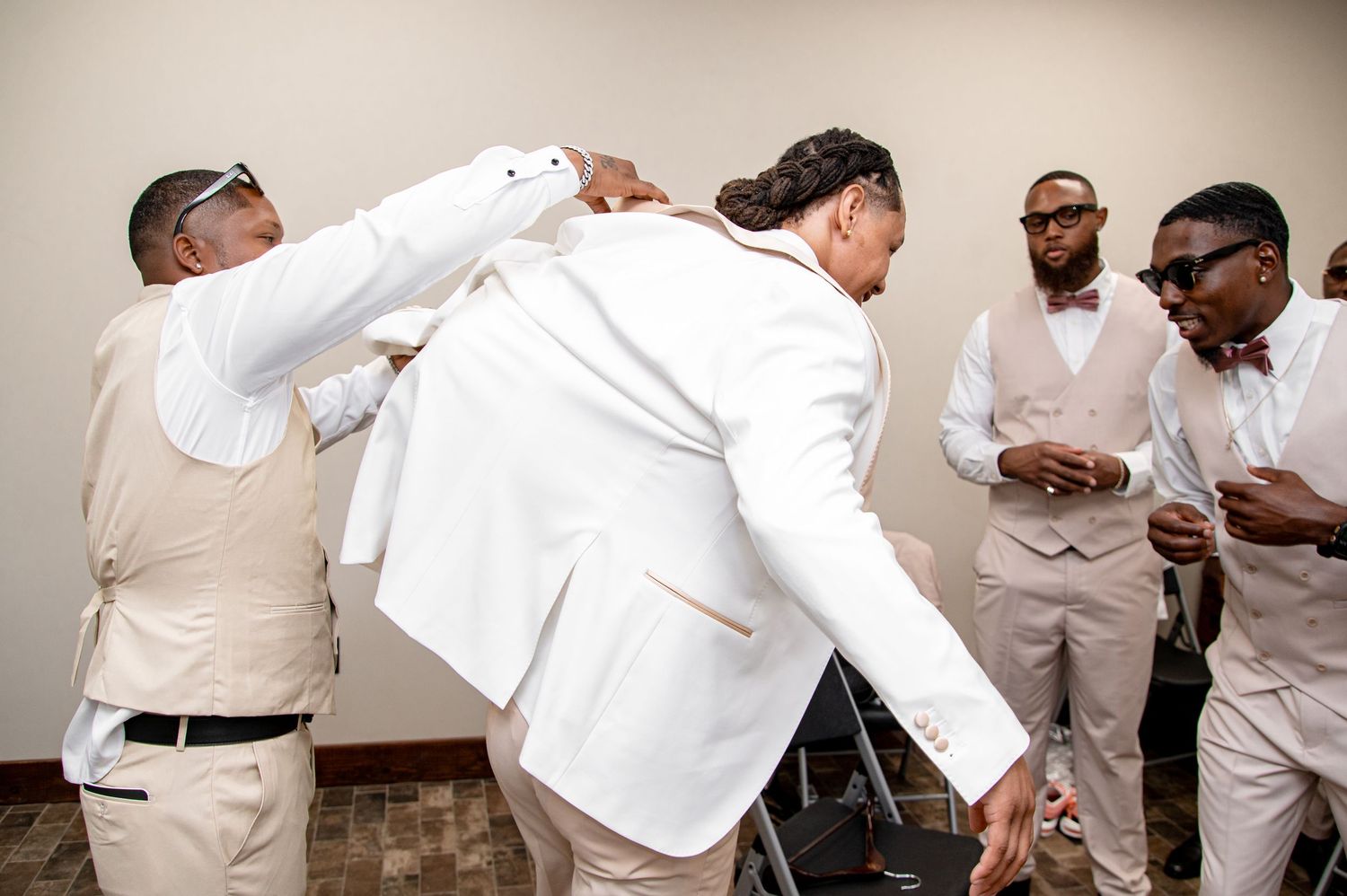 Groomsmen in beige vests help groom into white blazer while preparing for wedding ceremony.