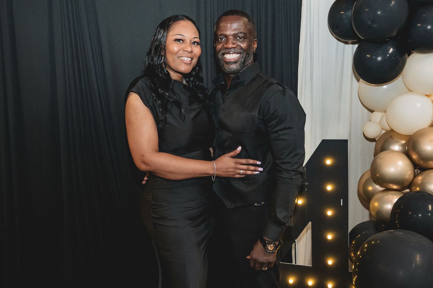 A couple in matching black formal attire smiles together at an event with gold and black balloon decorations.