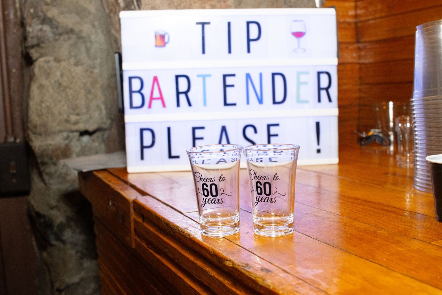 A handwritten 'TIP BARTENDER PLEASE!' sign sits on a wooden bar next to two empty shot glasses.