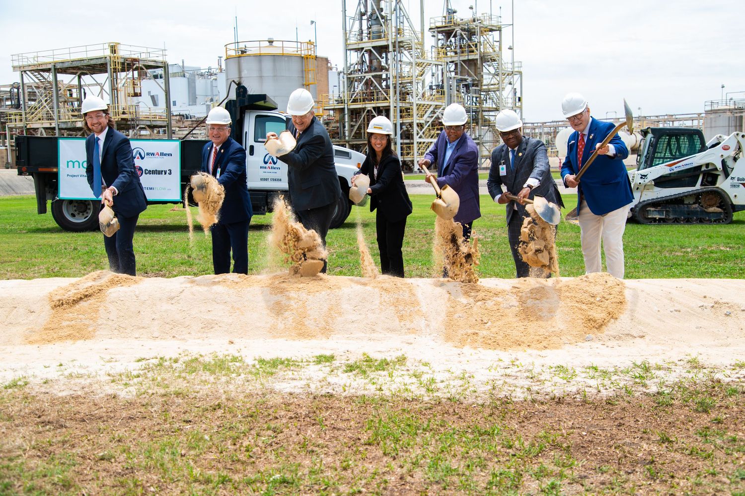 Group of executives in hard hats performing ceremonial groundbreaking at industrial facility construction site.