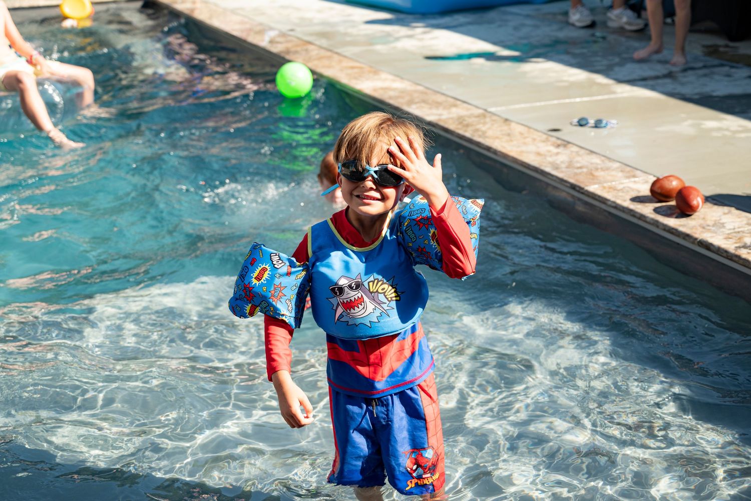 Young child in Spider-Man swimsuit and sunglasses playing in a sunny outdoor swimming pool.