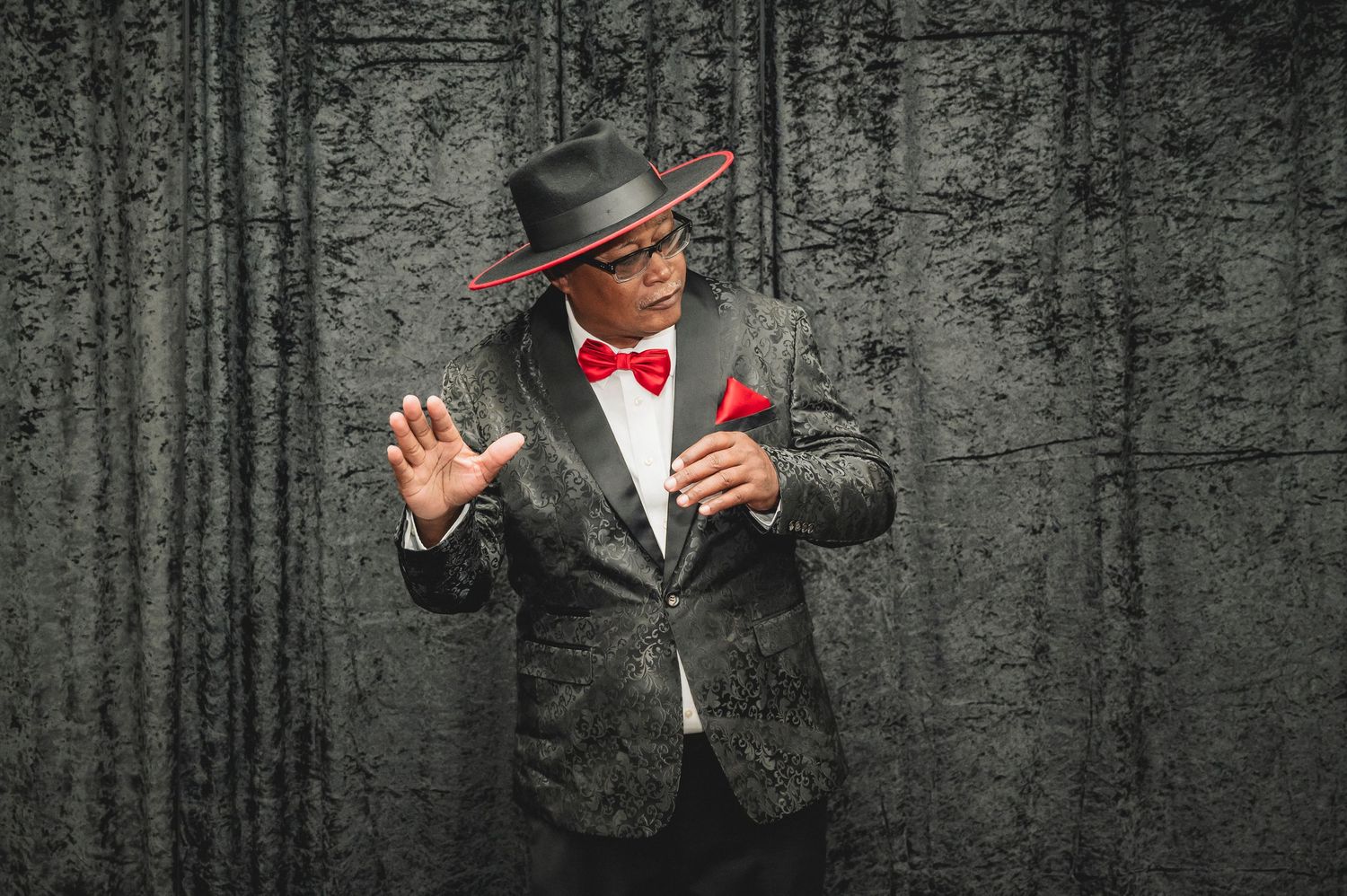 Person in black suit and hat stands against dramatic dark textured wall.