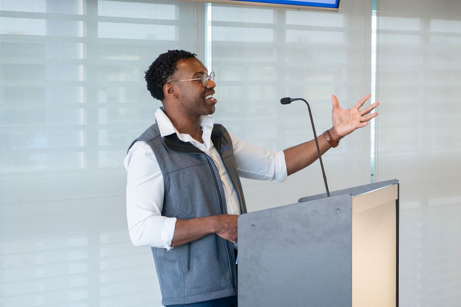 Black man in glasses and vest gestures while speaking at a podium with microphone during a presentation.