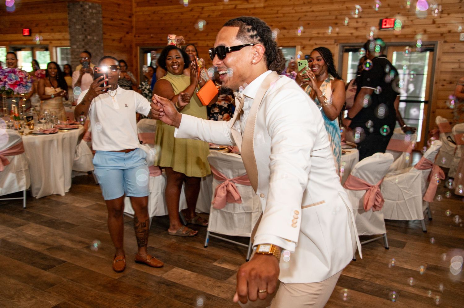 Groom in white suit dancing joyfully with guests at lively indoor wedding reception celebration.