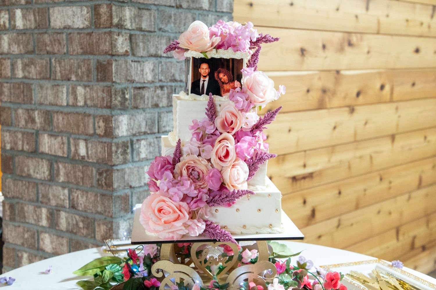 Four-tier white wedding cake adorned with pink and purple flowers and couple photo topper.