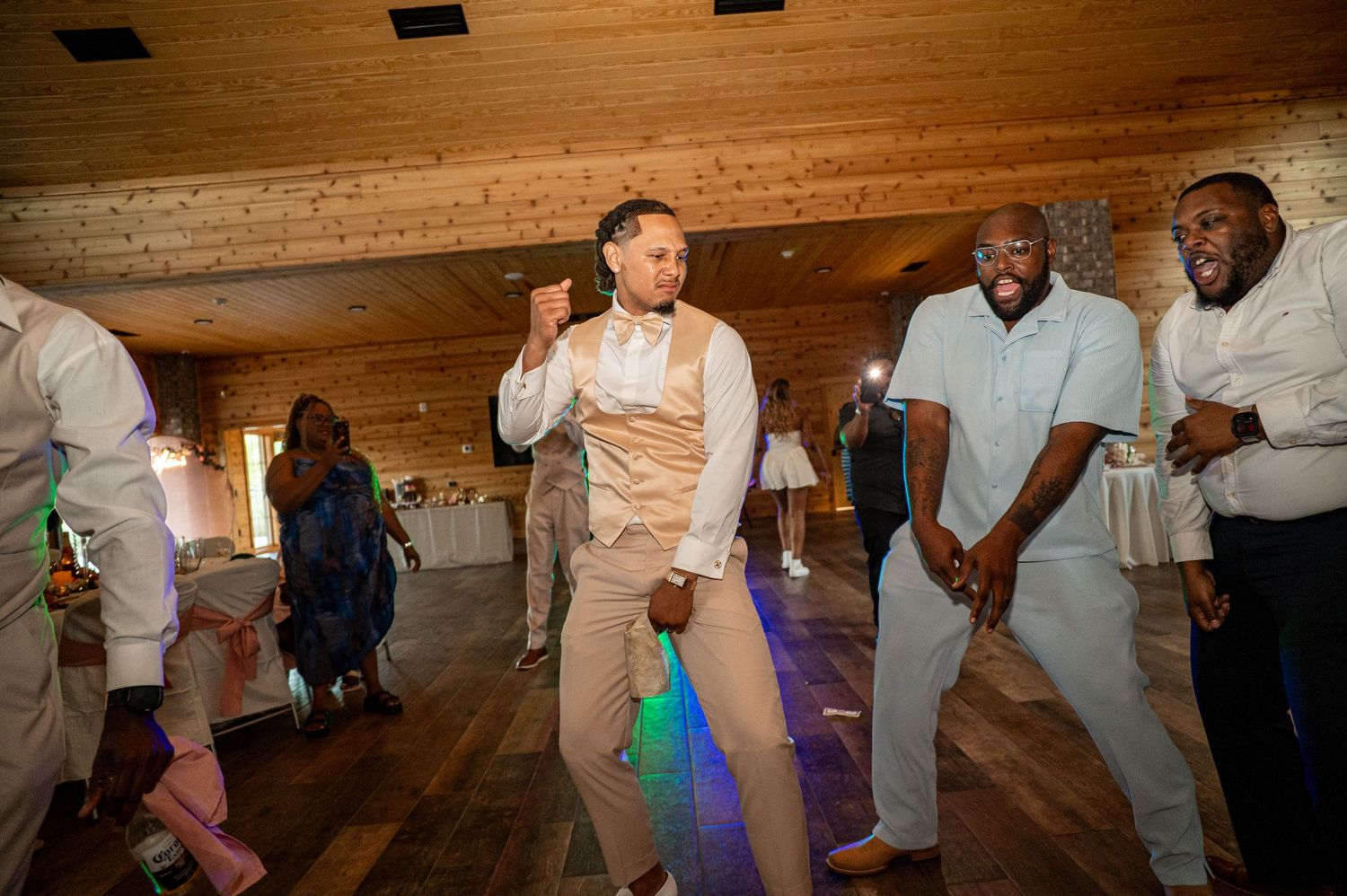 Groom in beige vest dances energetically with guests at lively wedding reception in rustic barn.