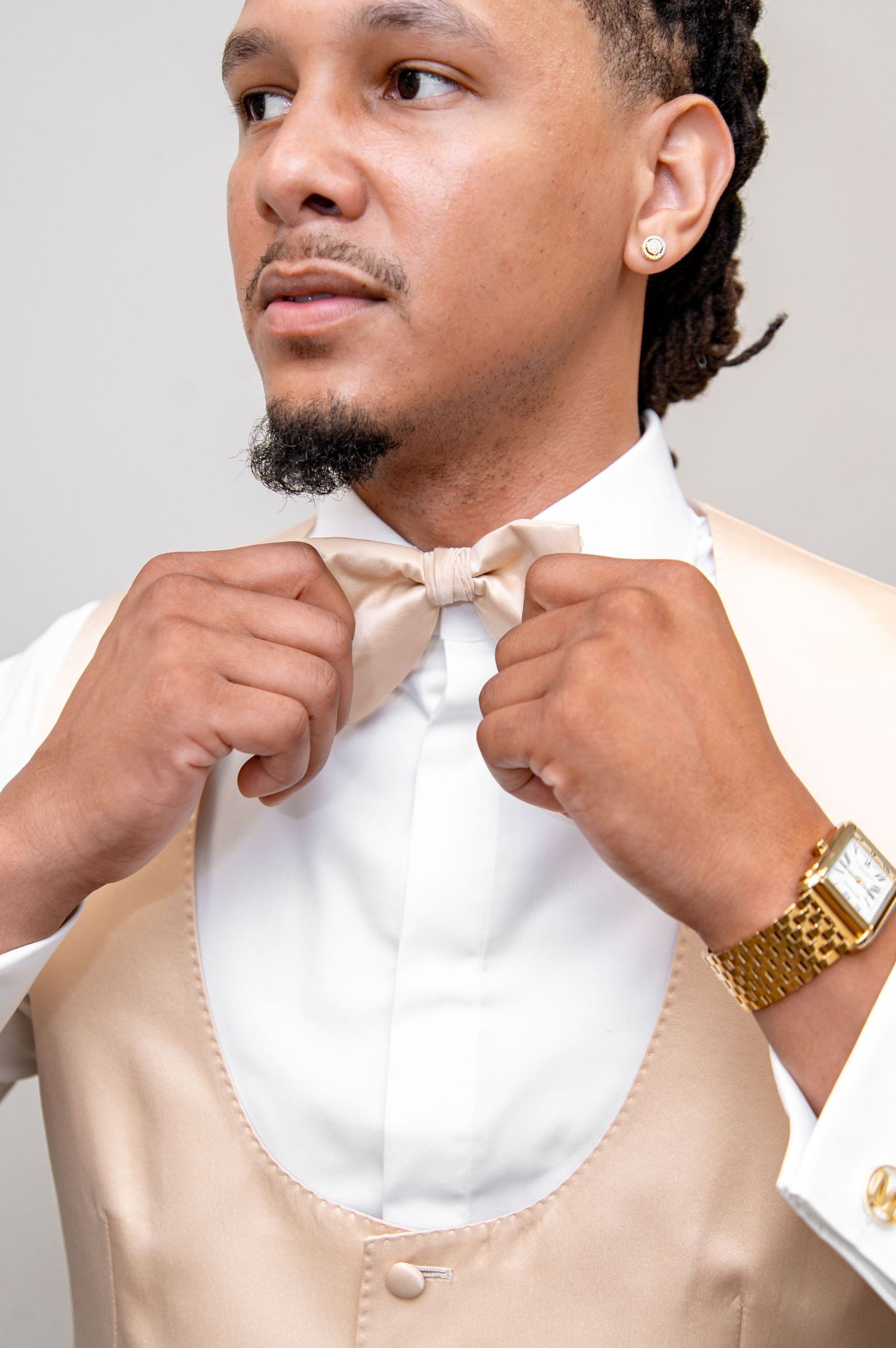 Man in beige tuxedo vest adjusting bow tie, wearing gold watch and earring.
