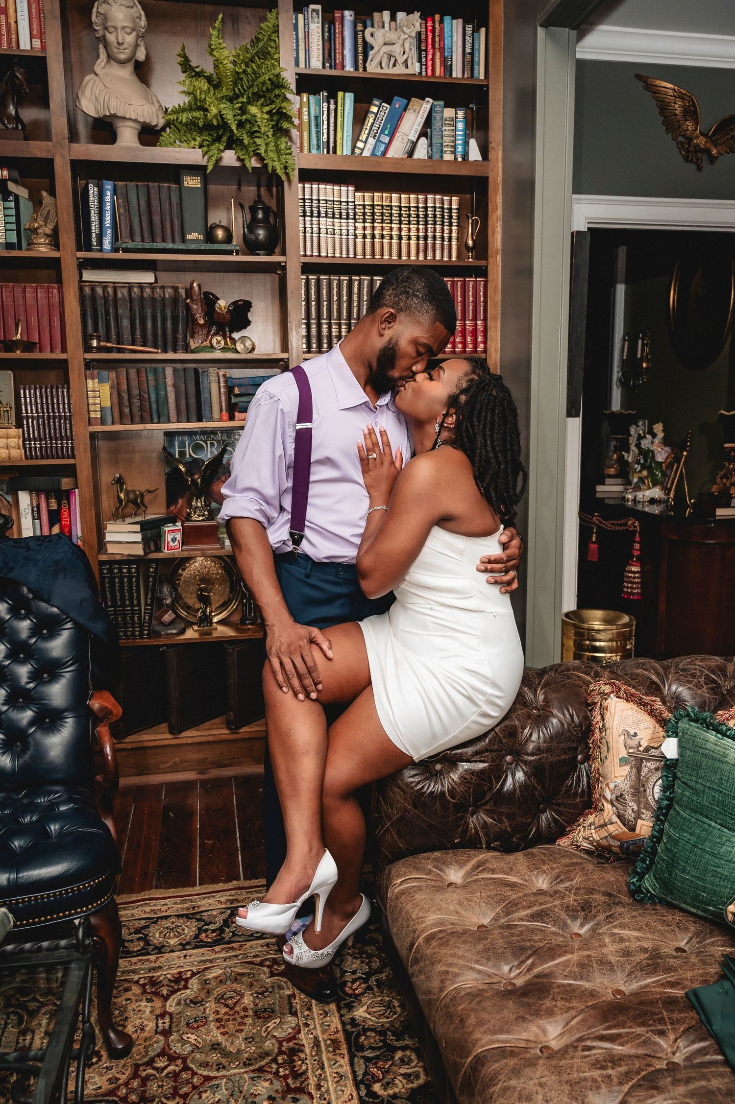A couple shares an intimate moment on a brown leather chaise in a home library setting with bookshelves in the background.