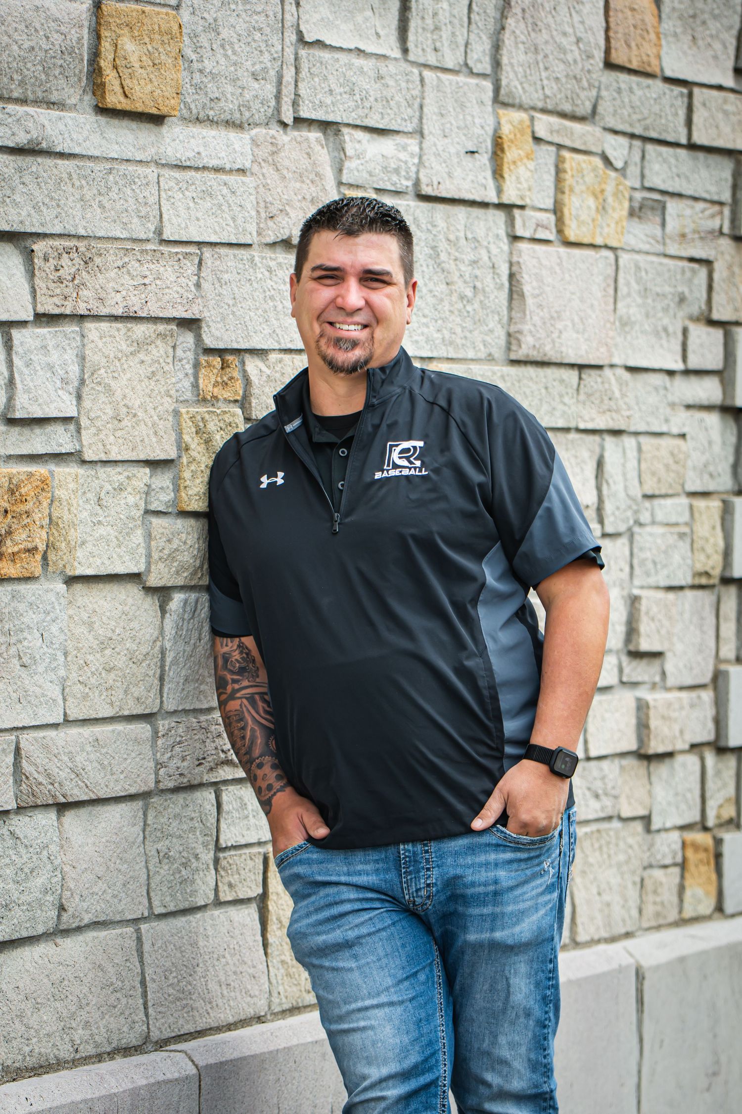 Man in black polo shirt poses against stone wall wearing jeans for casual headshots in Enumclaw, WA