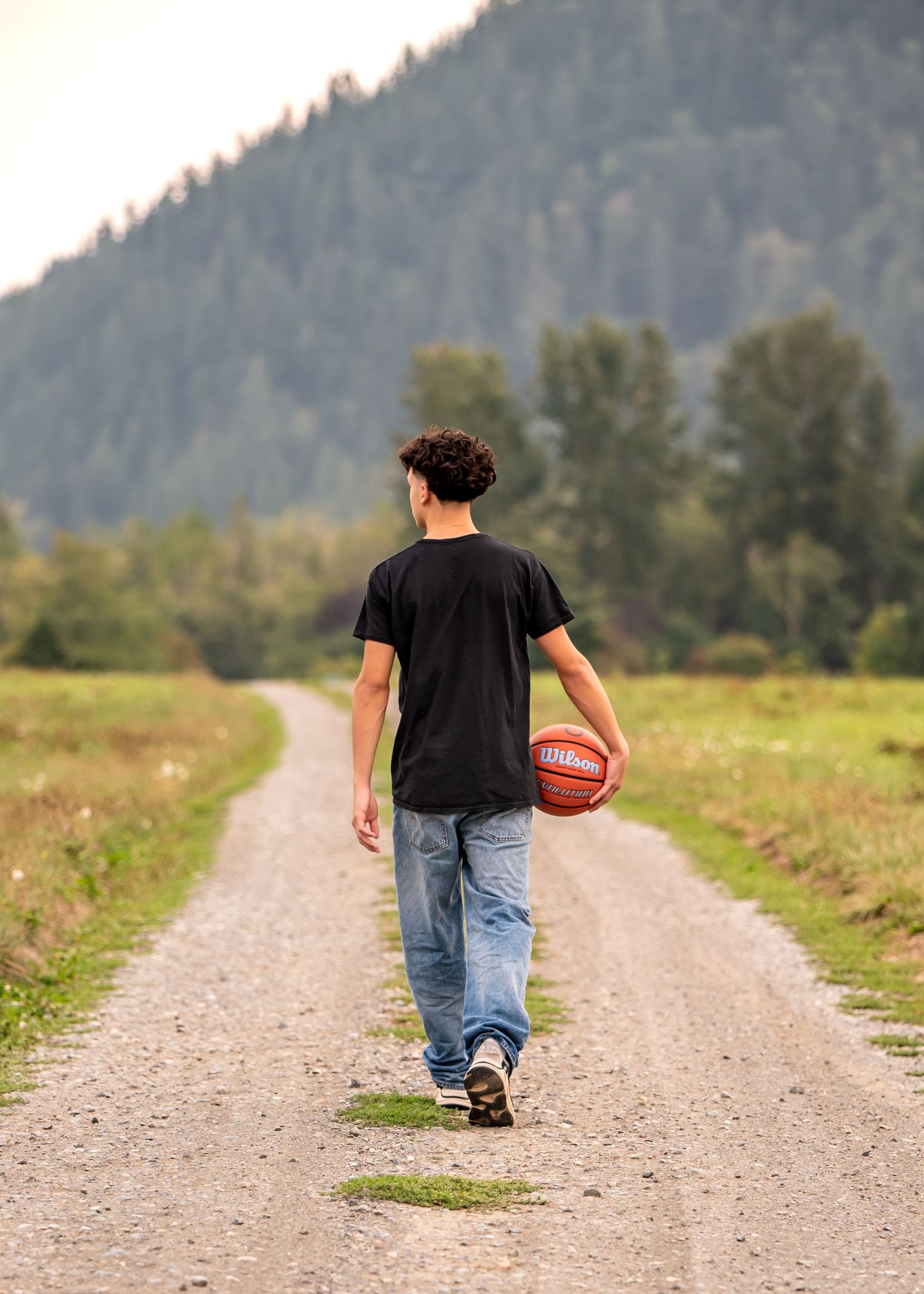 Tahoma high school senior walking alone on dirt path carrying basketball with mountains and trees in background in Enumclaw, WA