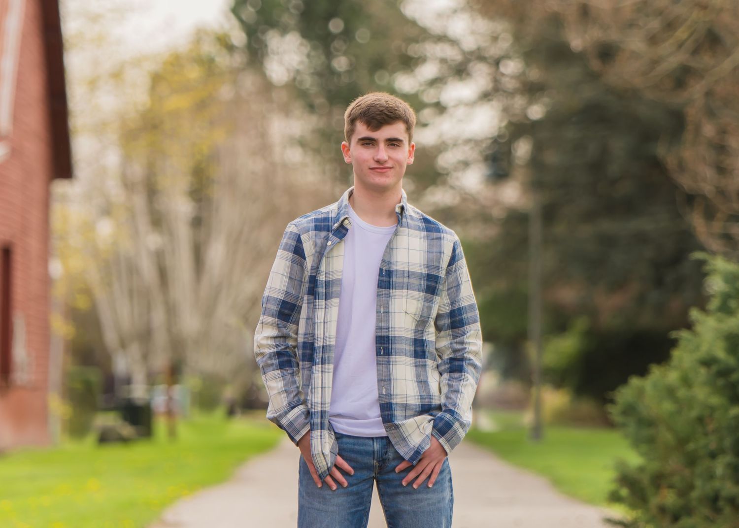 A HS senior in a plaid shirt and jeans stands on a paved path surrounded by green grass and trees during a senior session in Buckley, WA