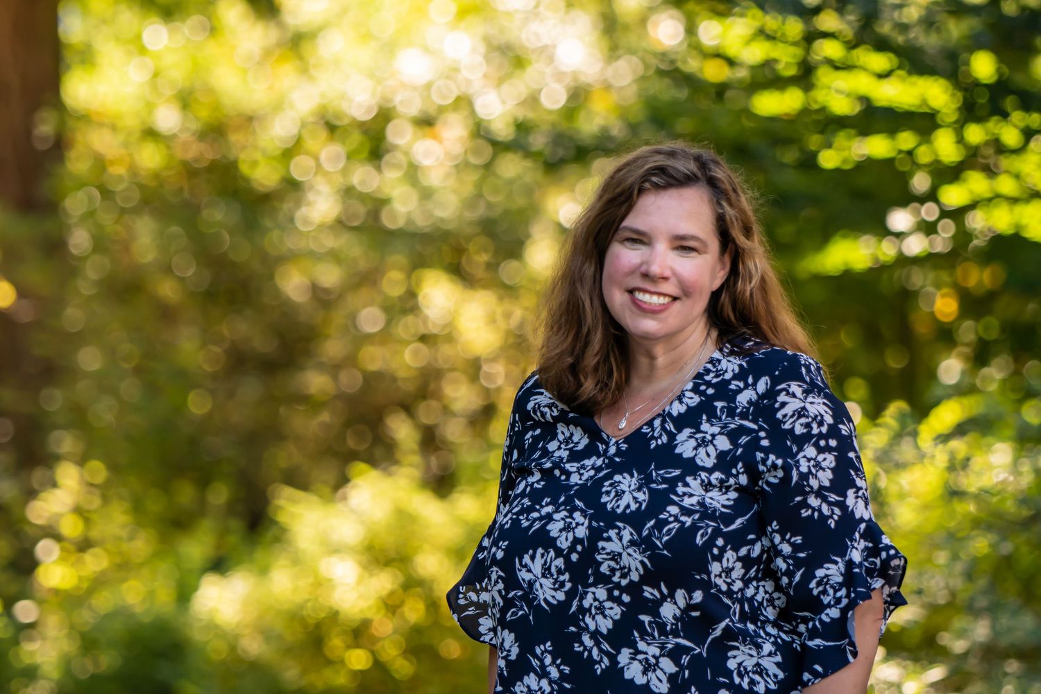 Woman in floral navy dress poses outdoors with sunlit trees in background.