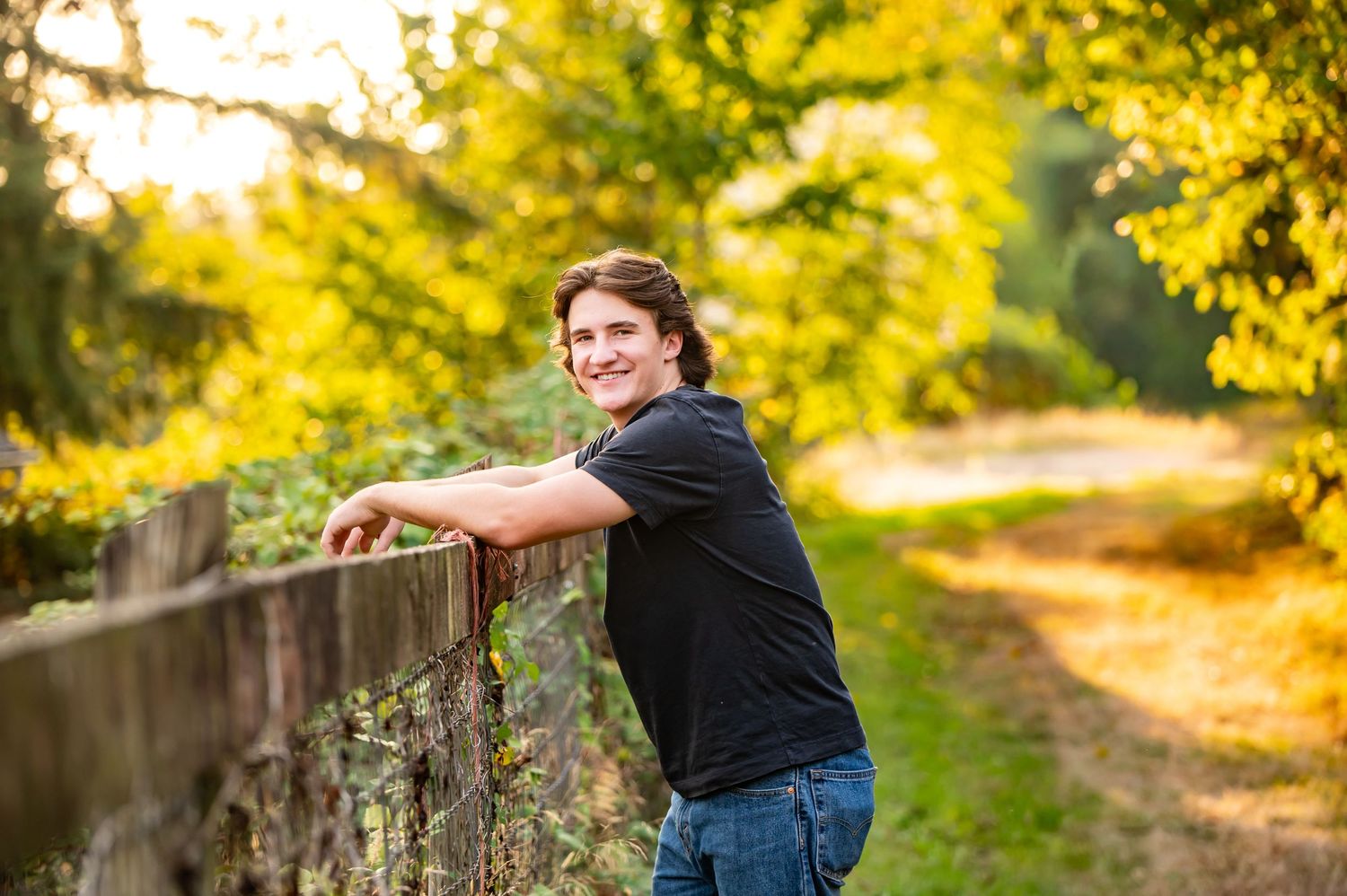 A Tahoma high school senior in black clothing poses along a rustic wooden fence at sunset in a lush autumn setting for senior photos