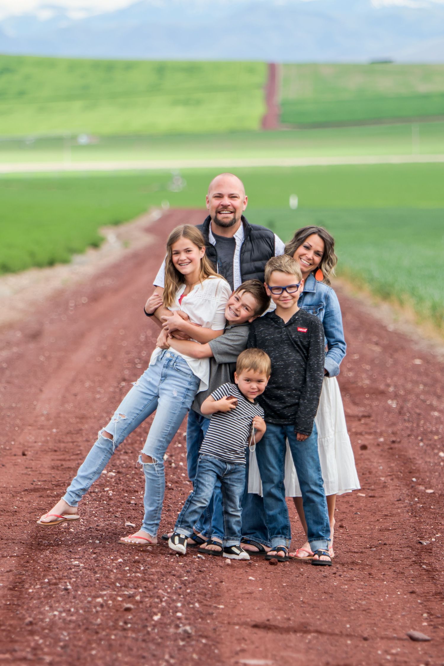 A playful family poses together in a sequence of photos on a dirt path surrounded by green fields.