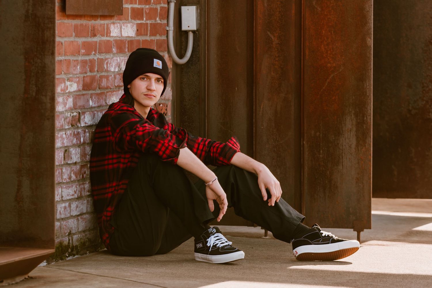 Person in red plaid shirt and black beanie sitting against brick wall for senior photos in Tacoma, WA