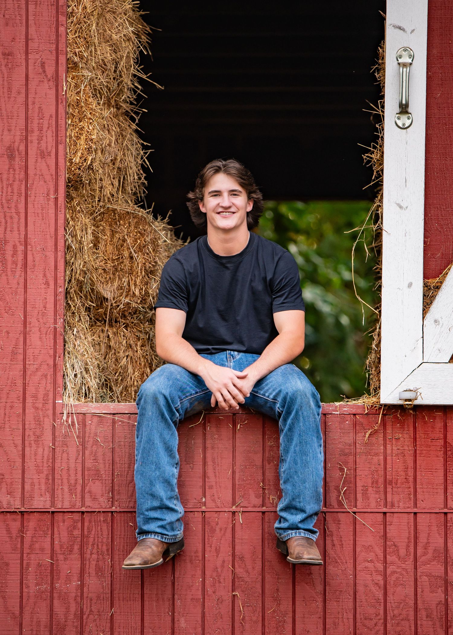 Tahoma High School senior sitting casually in openings of a rustic red barn with hay bales visible in the background during a senior shoot