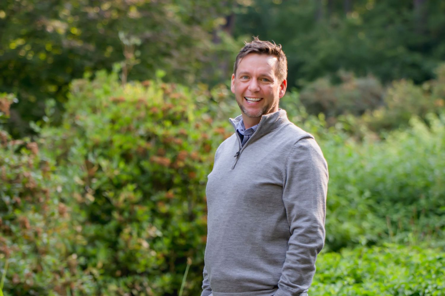 Man in grey quarter-zip sweater smiles in outdoor autumn setting posing during a corporate Headshot session in Maple Valley, WA