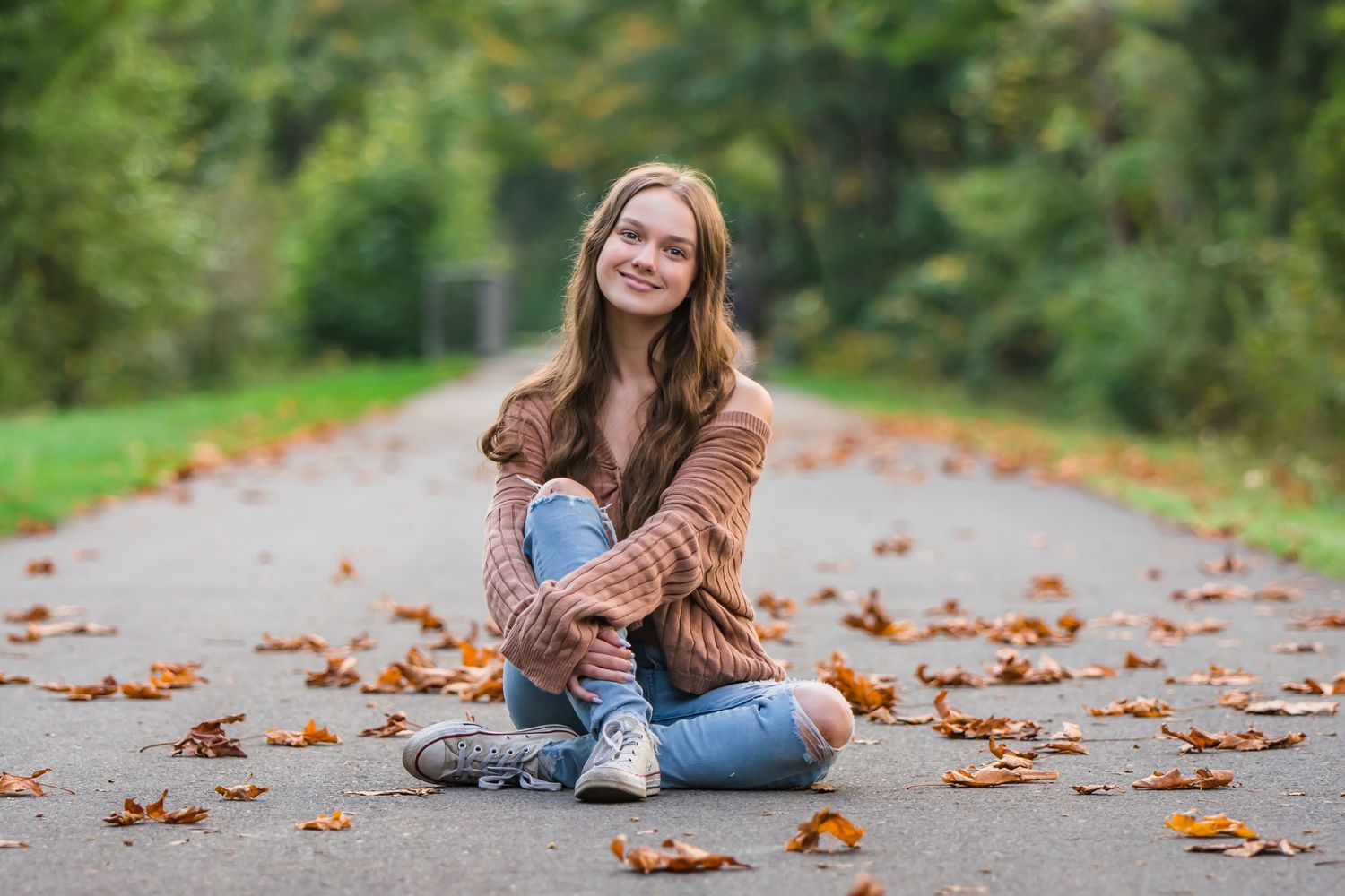 Kentlake Senior sitting on path covered in autumn leaves wearing brown sweater and ripped jeans during a senior photo shoot in Maple Valley