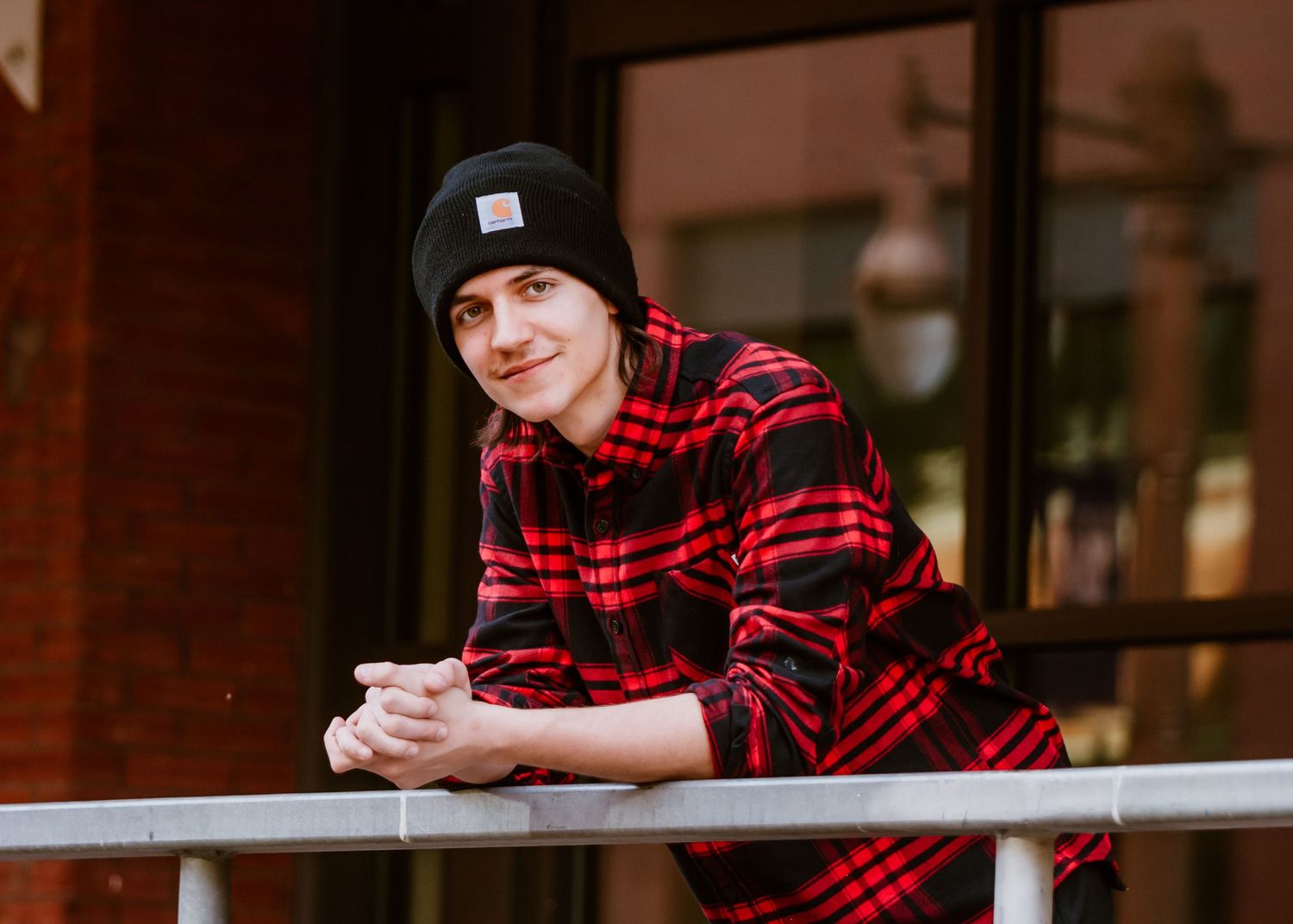 Tahoma senior in red plaid shirt relaxing on railing outside storefront during a senior photo session in Tacoma, WA