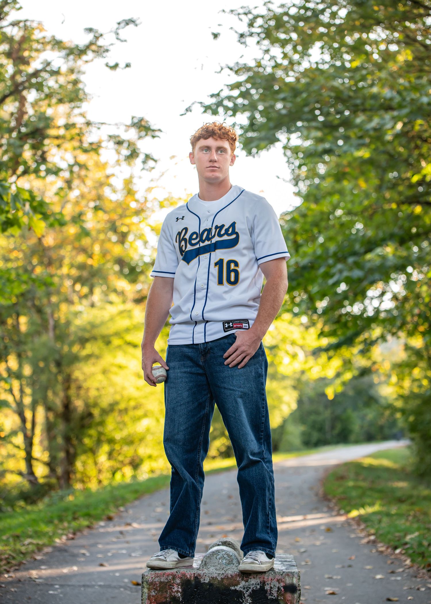 Tahoma Senior in baseball jersey standing on barrier along autumn path during senior photos