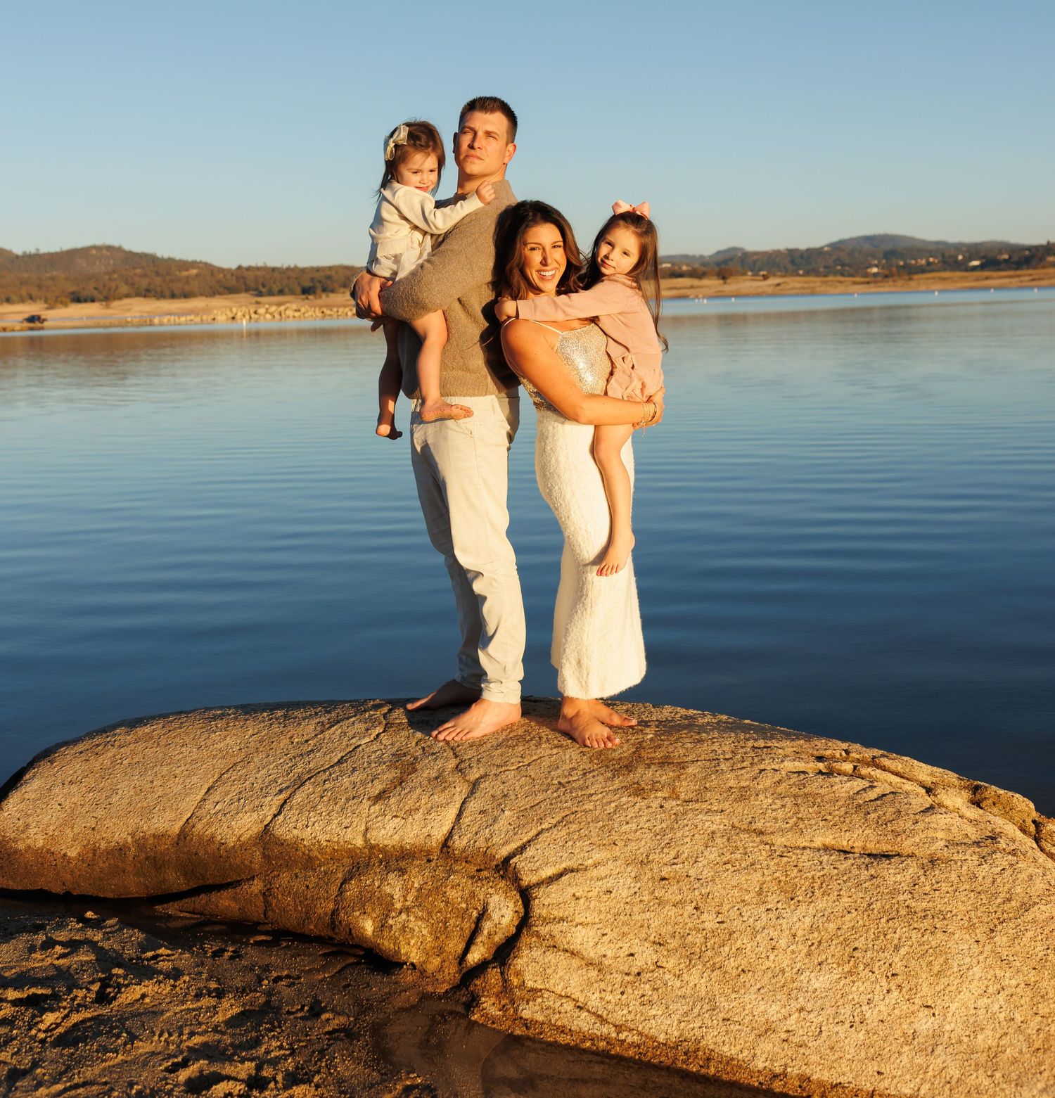 Family enjoying sunset together on large rock formation overlooking serene lake with mountains in background.