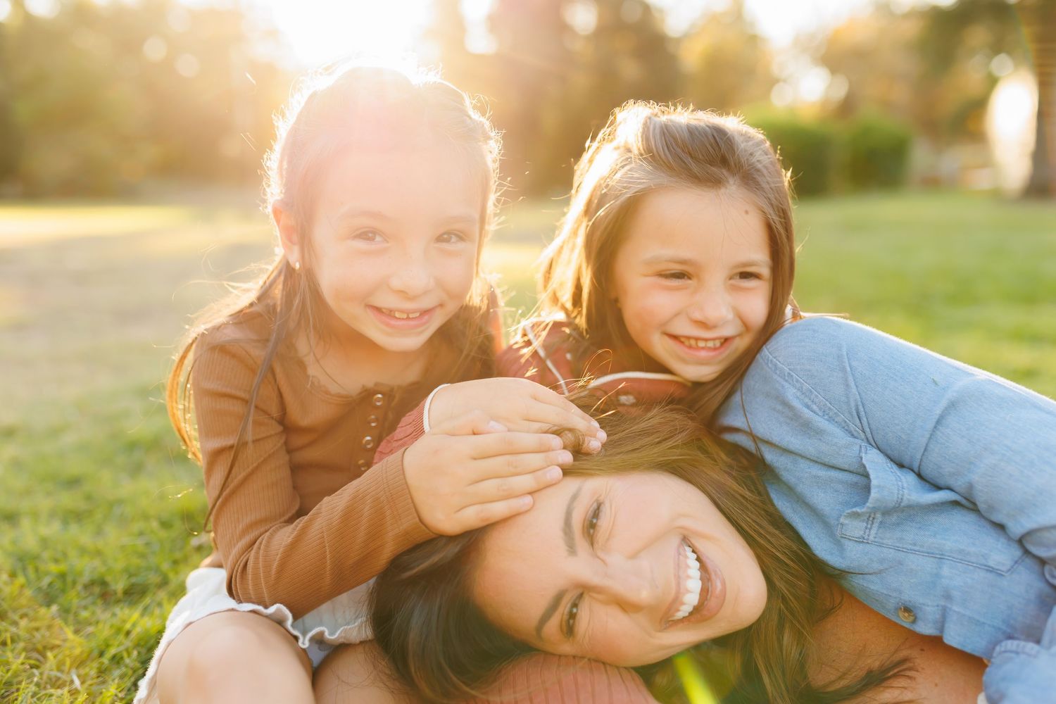 Two young girls joyfully hugging their smiling mother outdoors in a sunlit park during golden hour.