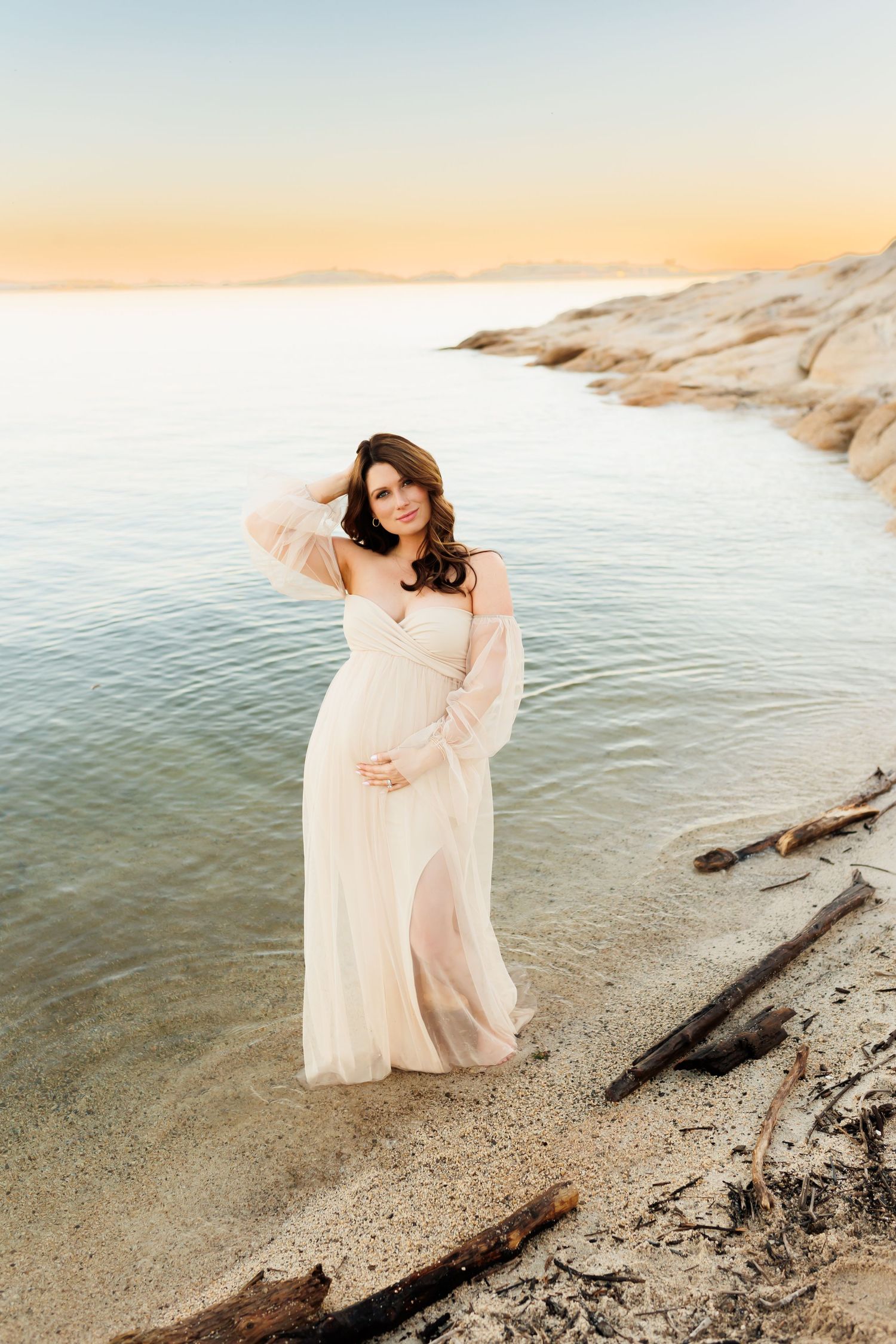 Woman in flowing white dress stands gracefully by the water's edge during sunset at a rocky beach.