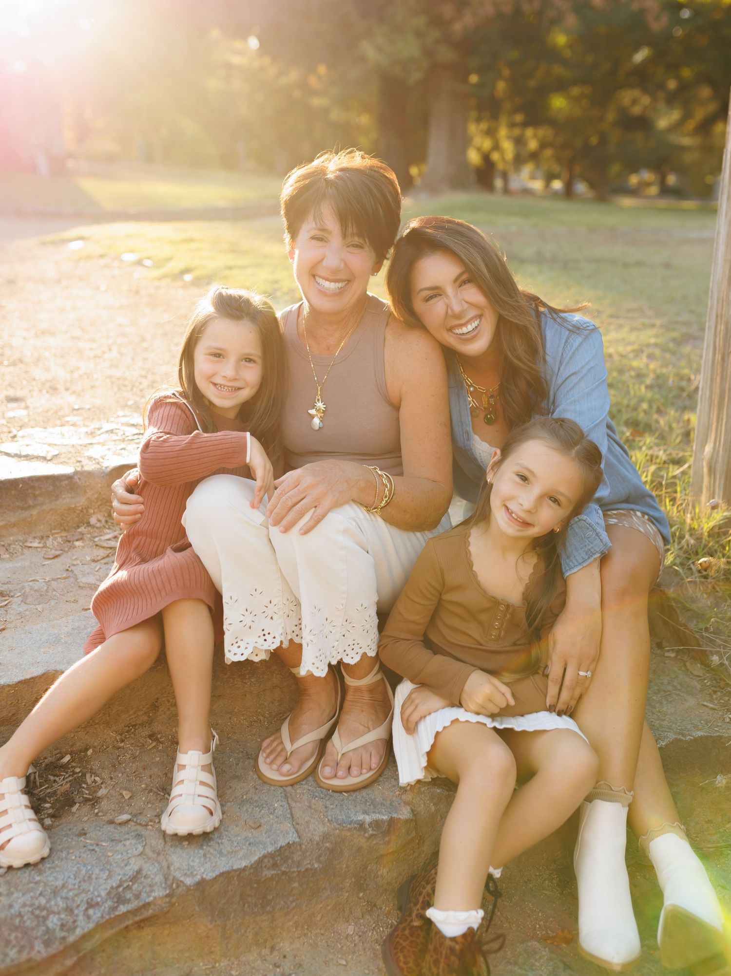Four generations of women smile together outdoors in warm golden sunlight, sitting on stone steps in a park.