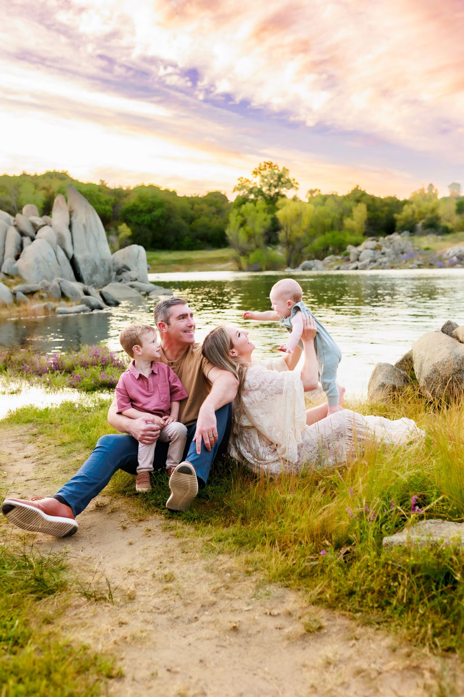 Family lounges on rocky shoreline at sunset near calm water.