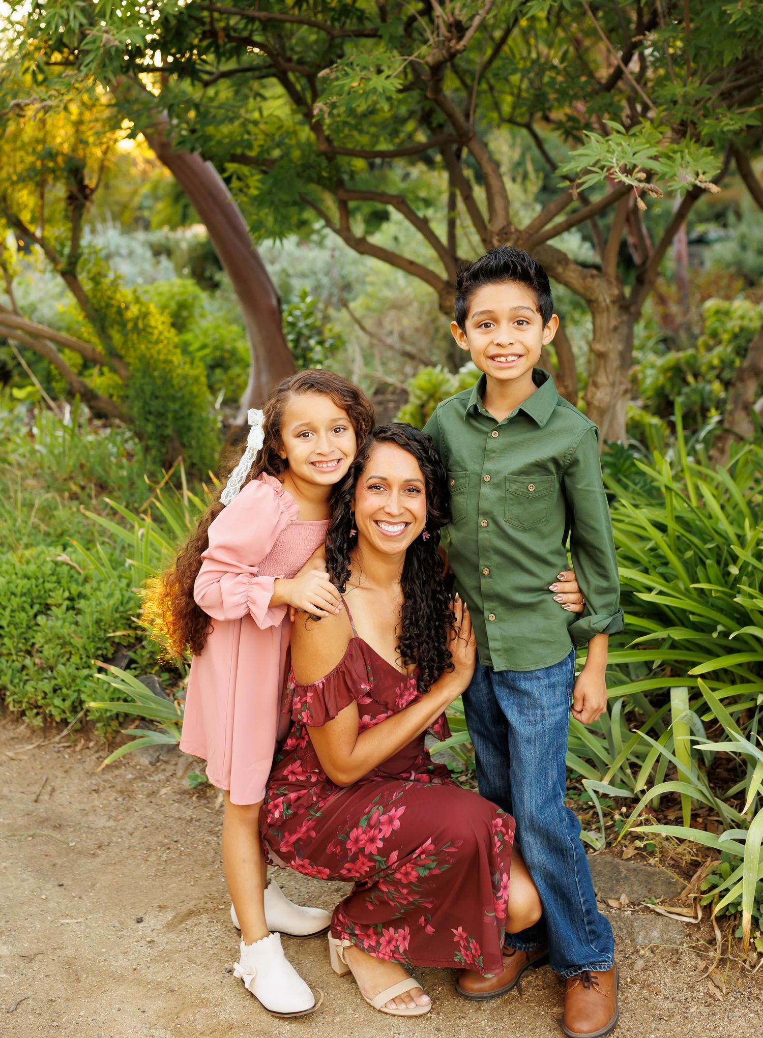 A smiling mother crouches with her two young children in a lush garden, wearing a floral dress for a family portrait.