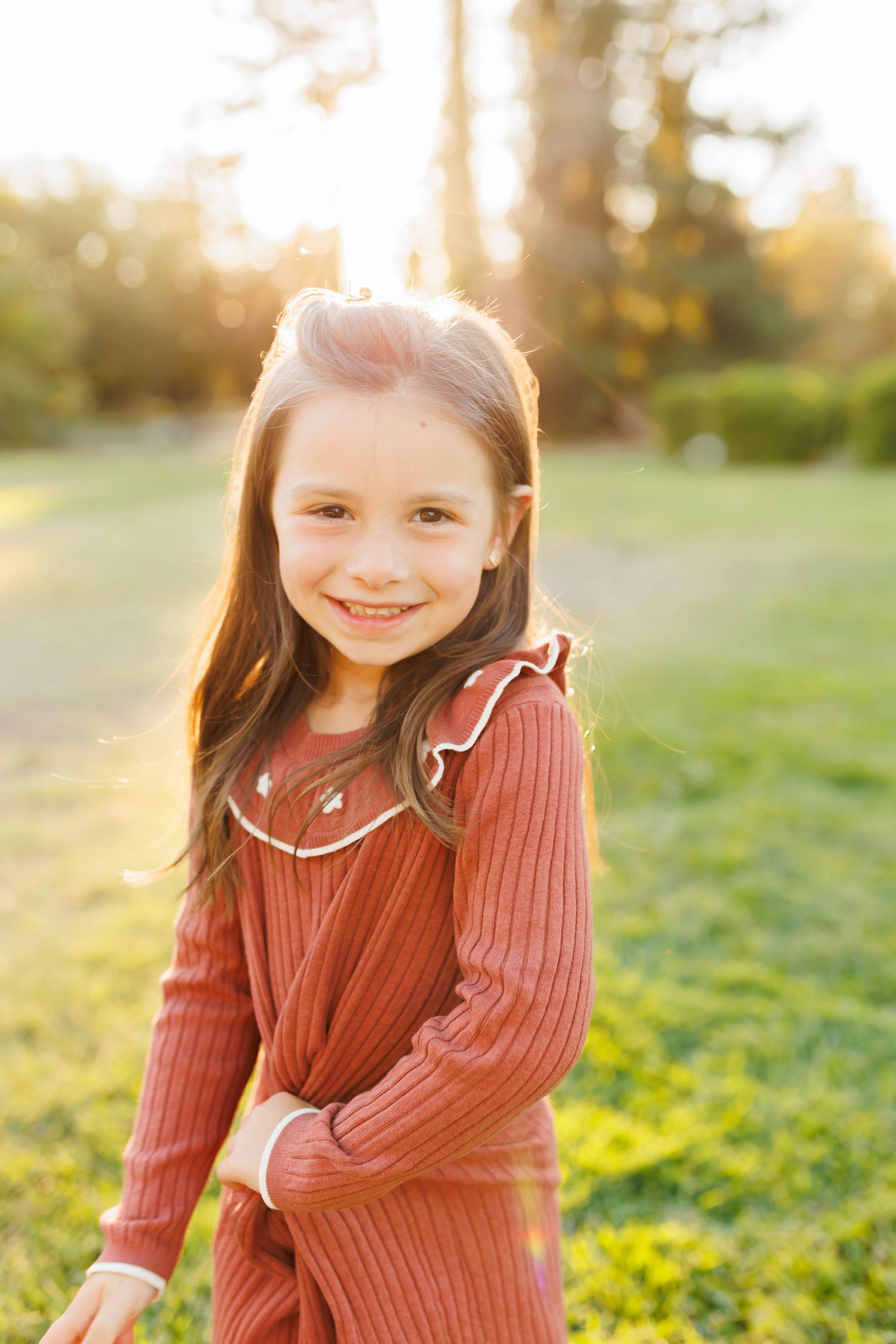A smiling young girl in a red ribbed dress poses outdoors in golden hour sunlight with green grass and trees behind her.