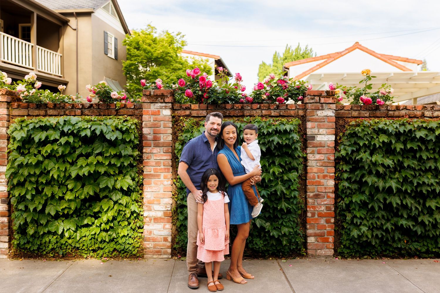 Family of four posing in front of a brick wall covered in lush ivy and pink roses outside a home.