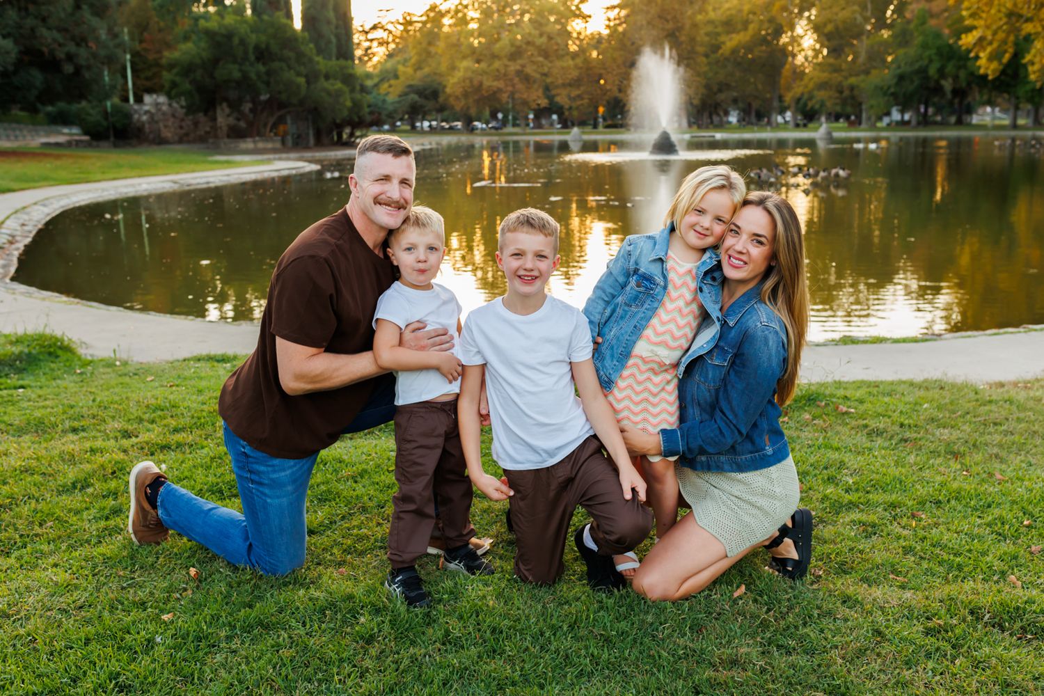A family poses together on grass near a pond with a fountain at sunset in a park setting.