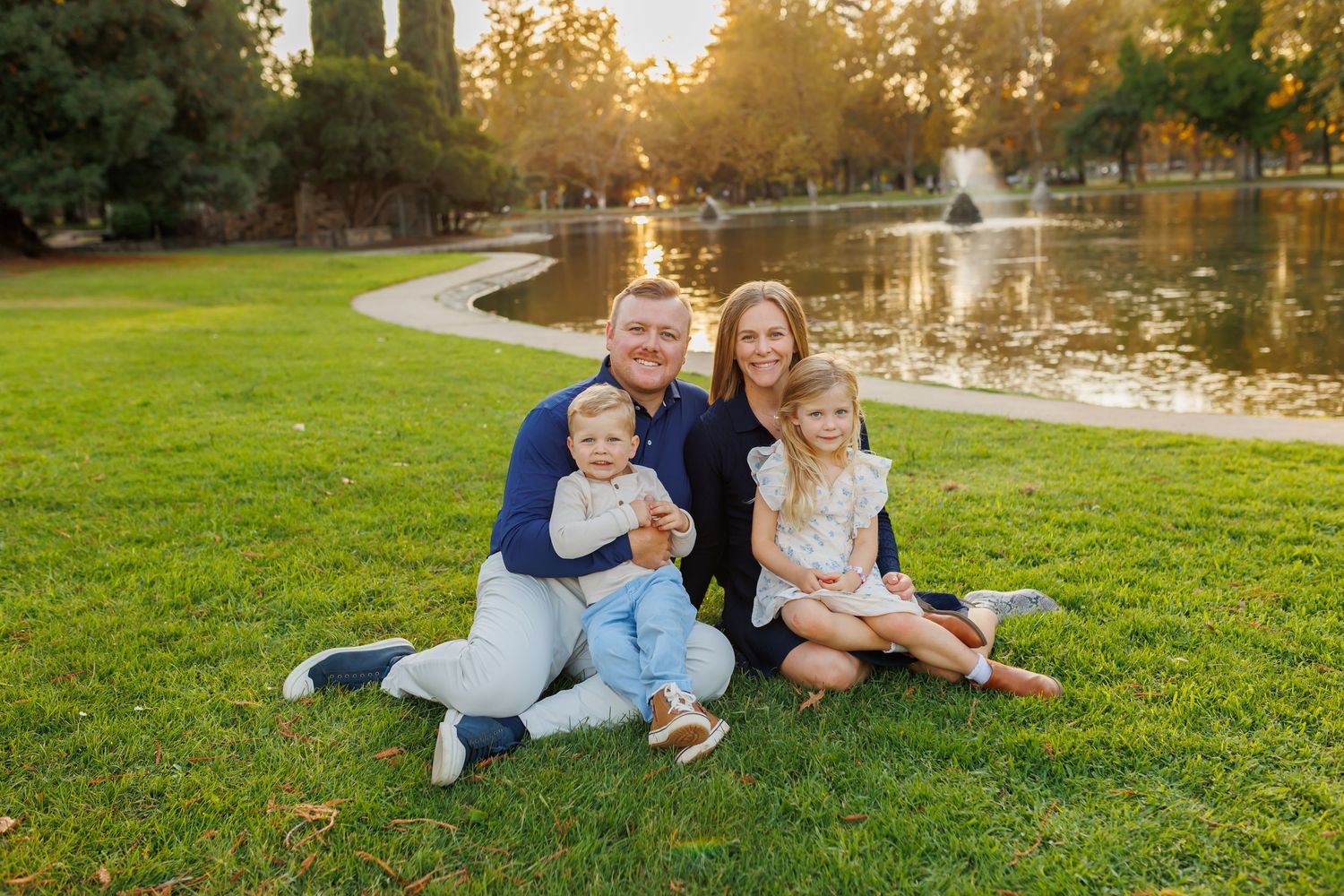 Family of four sitting on green grass by a pond at golden hour in a sunny park setting.