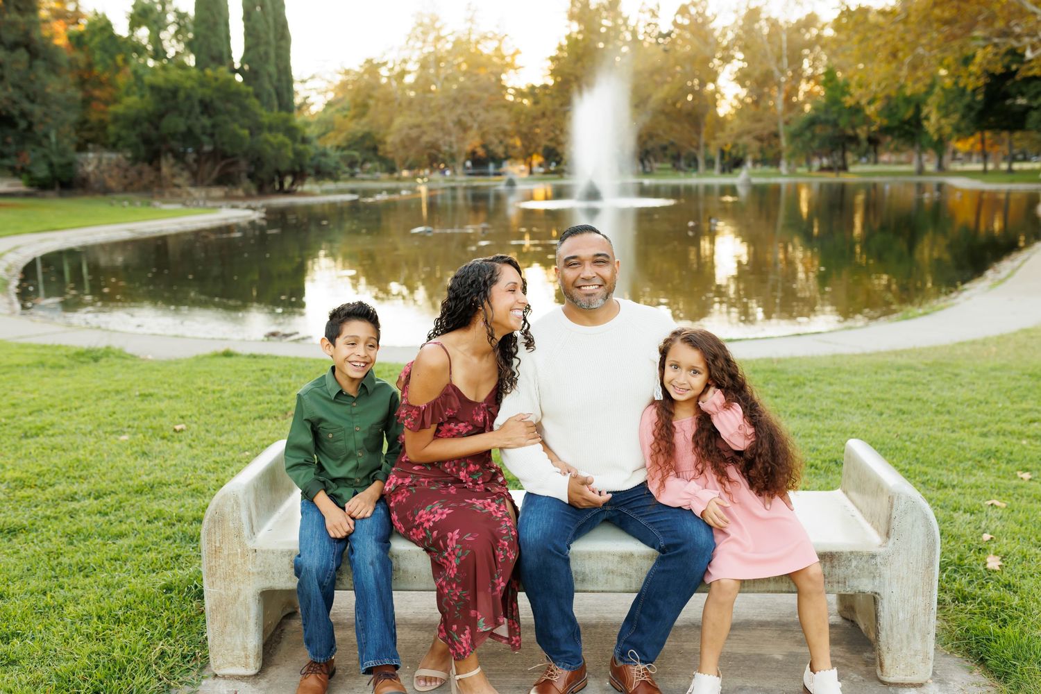 Happy family of four sitting on a stone bench by a pond with a fountain in a lush green park.