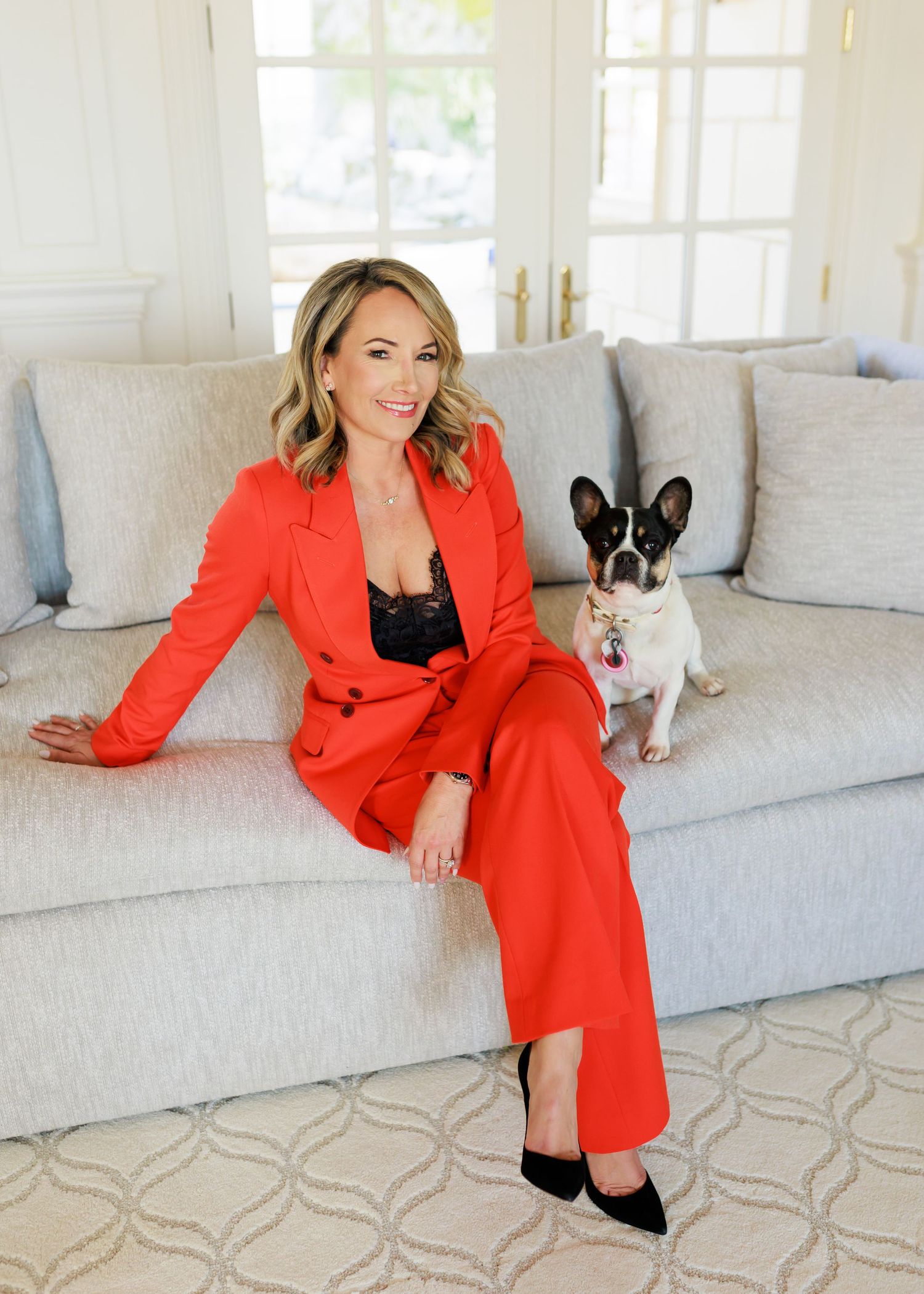 Lifestyle photoshoot with red blazer outfit and small dog on grey couch.