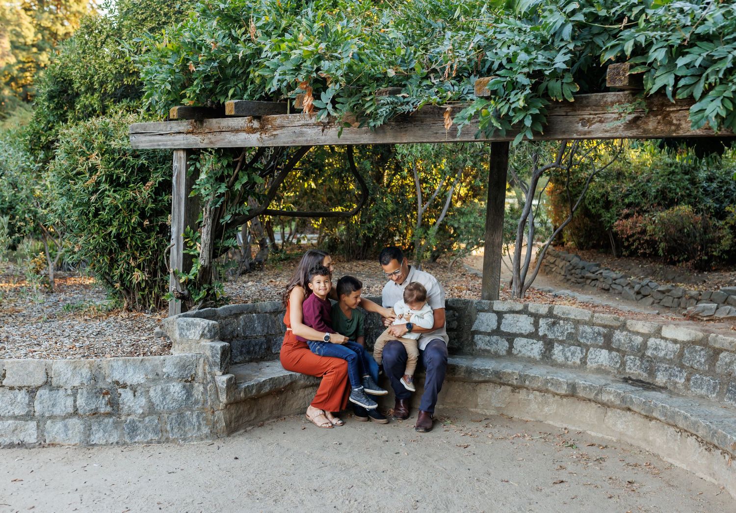 Family relaxing on stone wall in garden setting during sunset photo session.