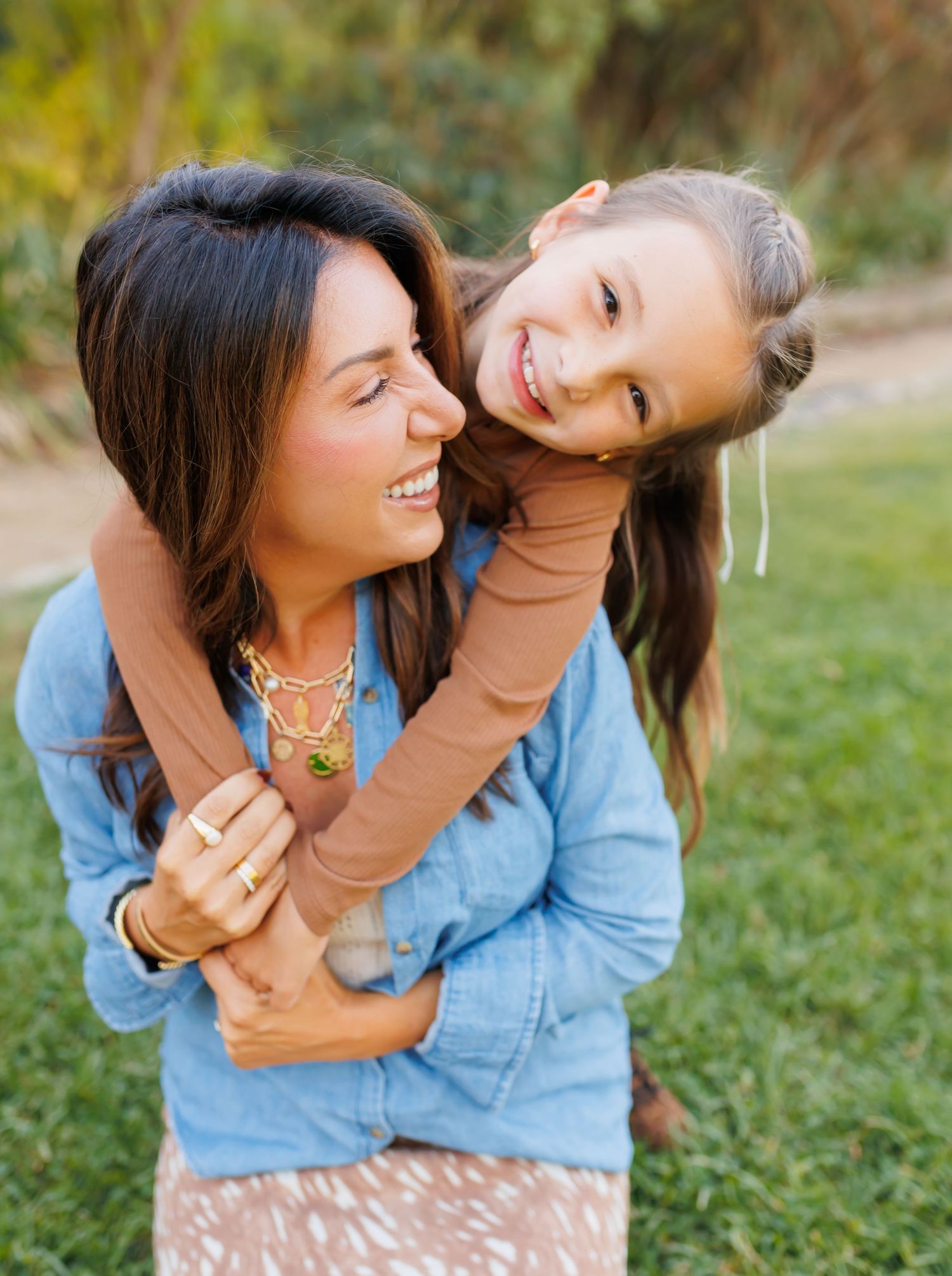 A smiling woman in a denim jacket carries a laughing young girl on her back outdoors on green grass.