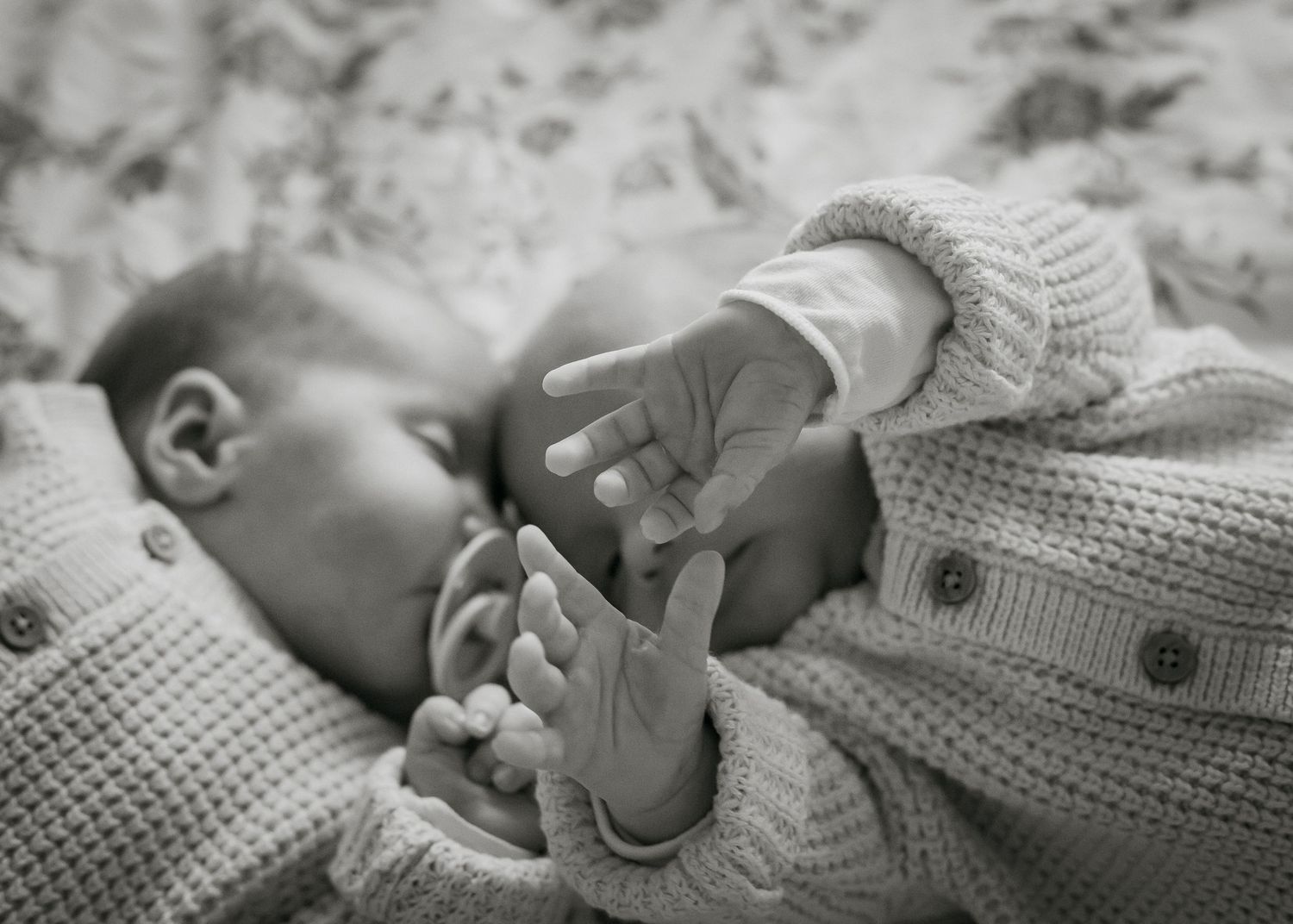 Black and white. Two newborn twins. one is sleeping and the other has his hands up in the air. Focus is on the hands of the awake twin.