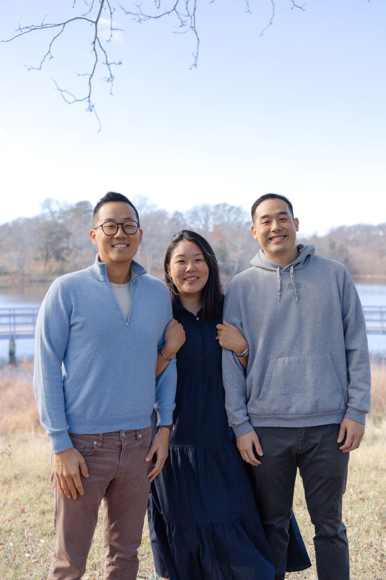 Three smiling friends pose outdoors near a river on a sunny winter day, wearing casual blue and gray outfits.
