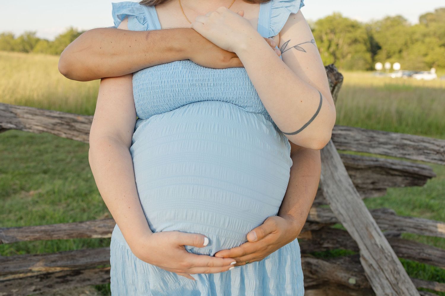 Pregnant woman in blue dress outdoors, partner embracing her from behind near rustic wooden fence in green field.
