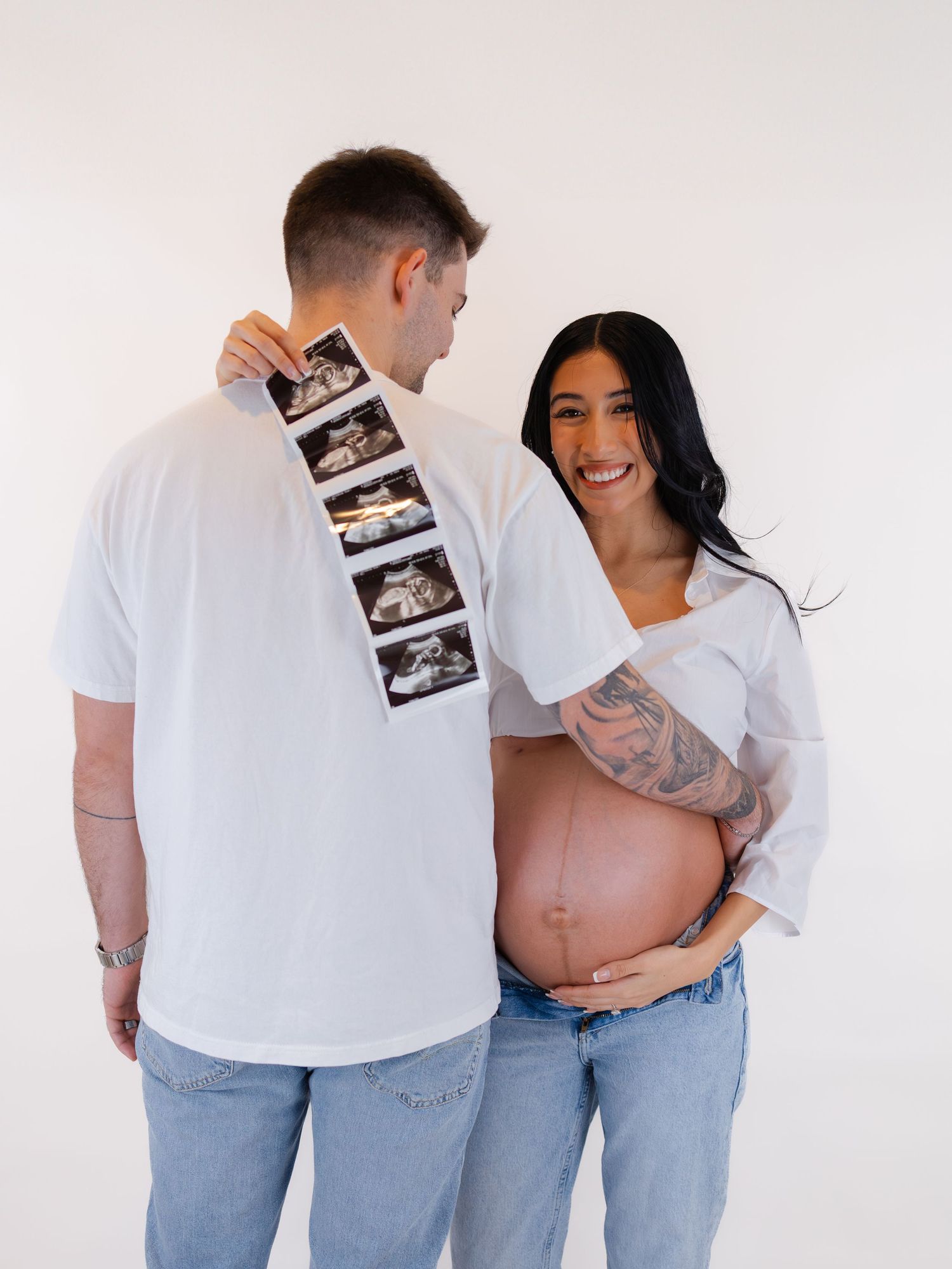 Smiling pregnant woman and partner holding ultrasound photos, maternity shoot in white studio.