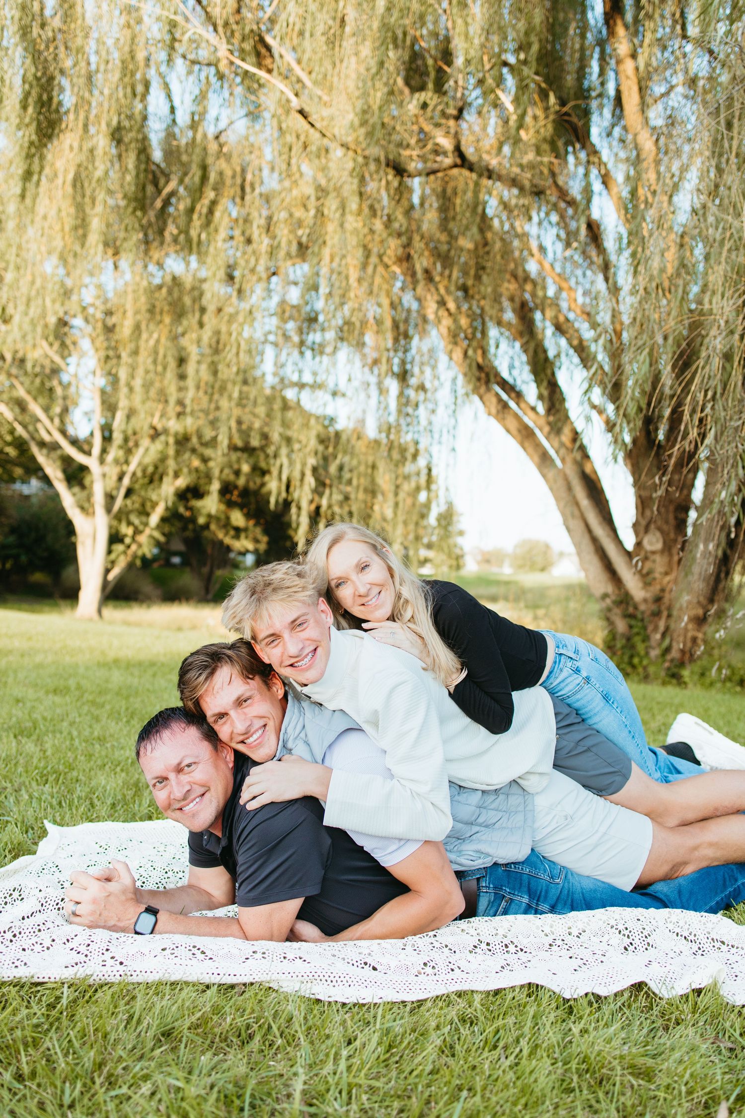 Happy family of four lying on a blanket outdoors, stacking on each other near a willow tree on a sunny day.