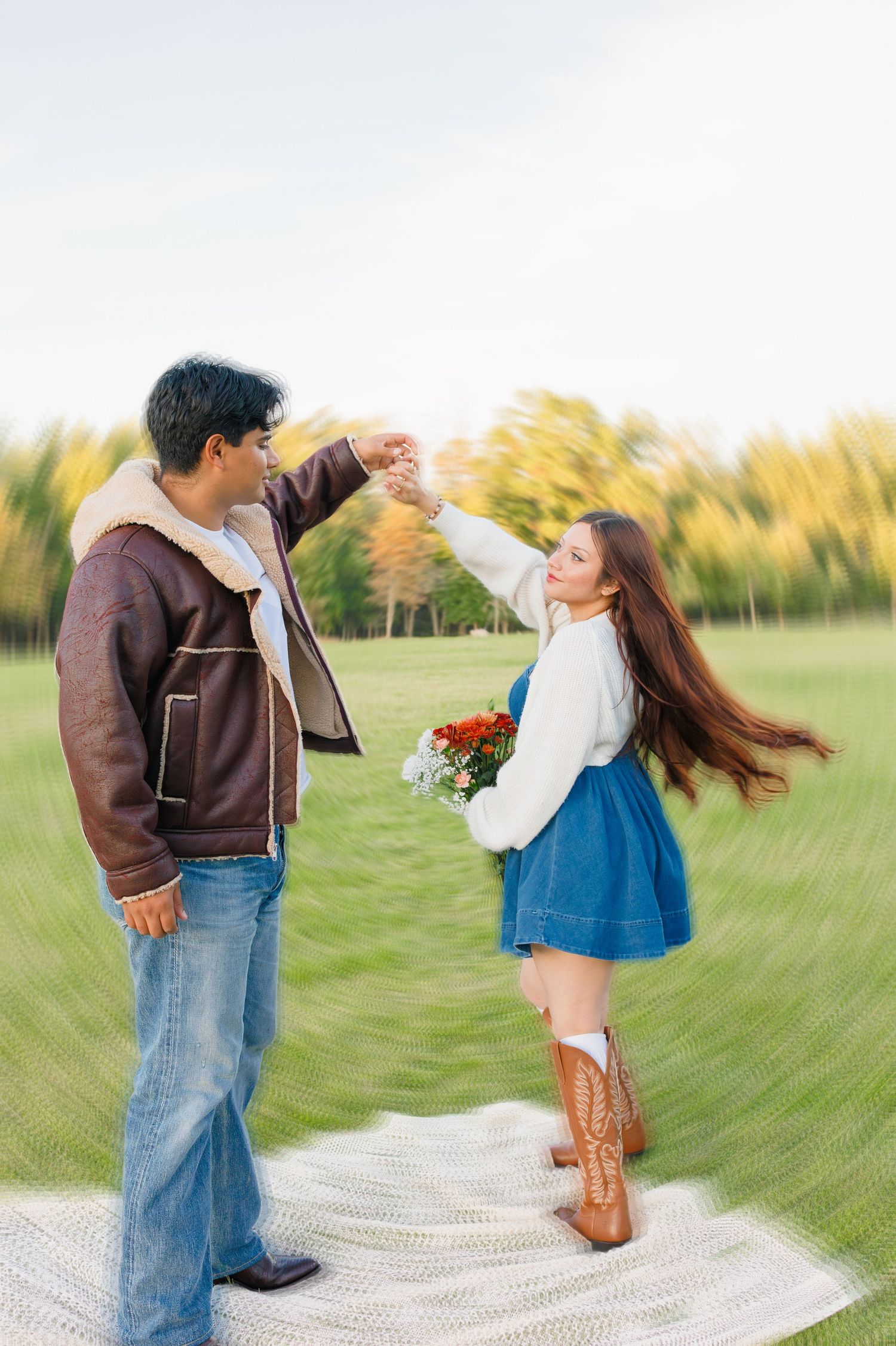 Couple dancing outdoors on a blanket in a green field, woman in blue dress and cowboy boots holding flowers.