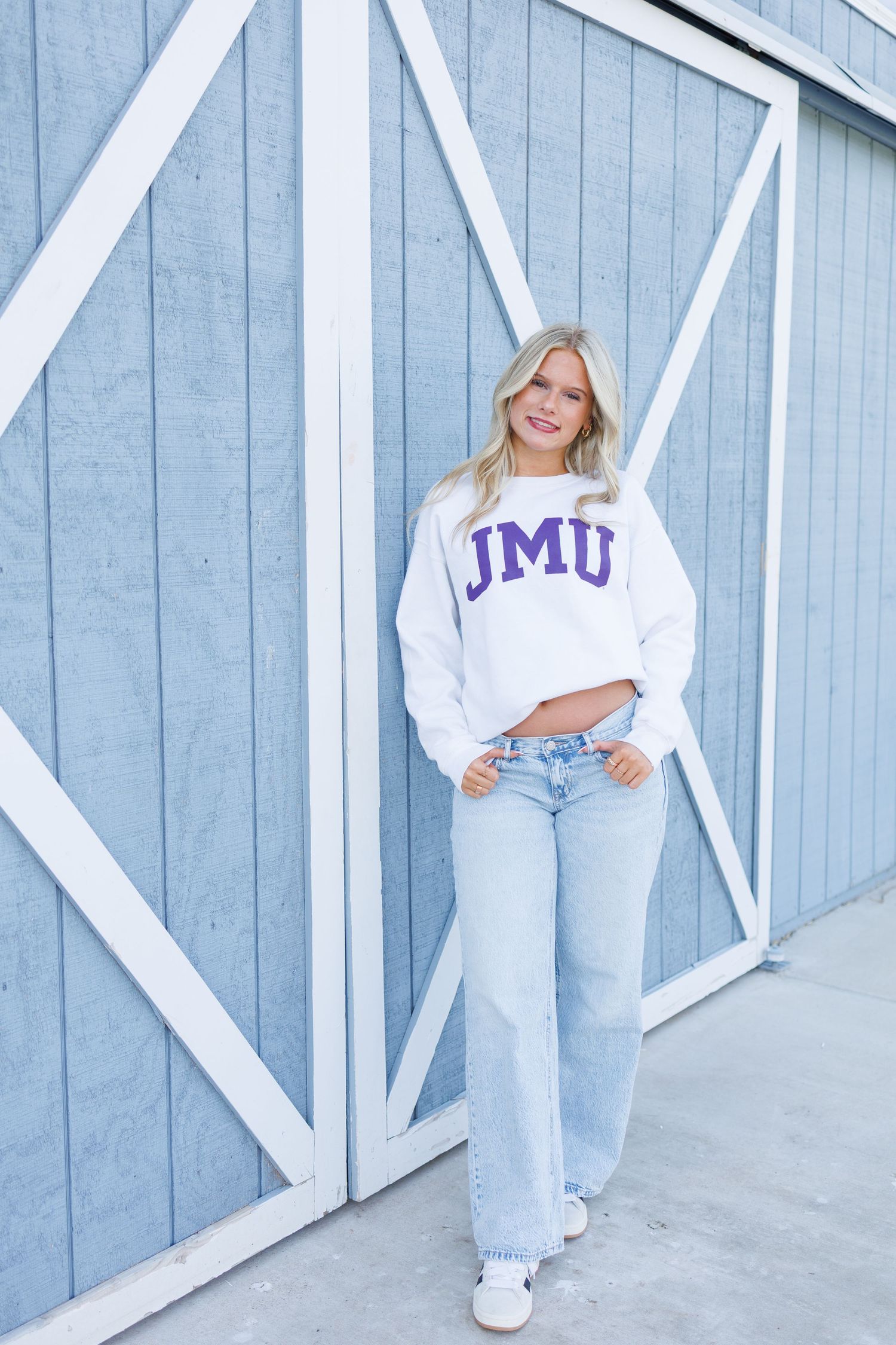 Woman wearing a white JMU cropped sweatshirt and light wash jeans, posing against a blue barn door.