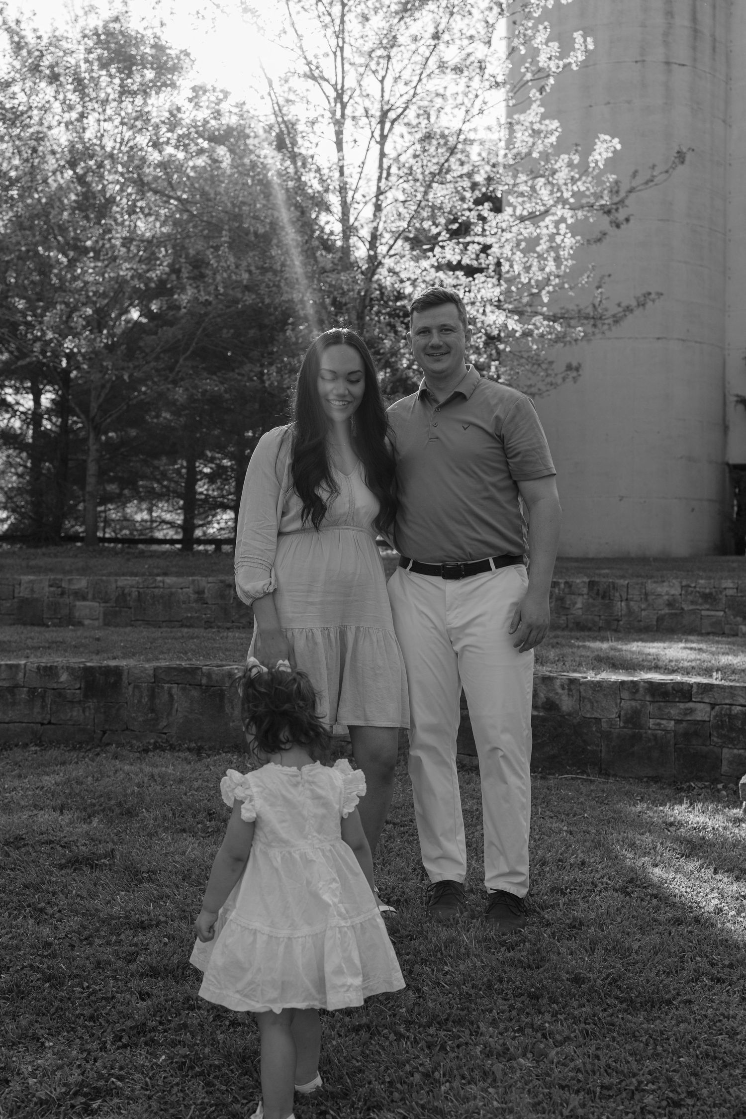Black and white family photo of a couple with young daughter in white dress standing on grass near stone steps and trees.