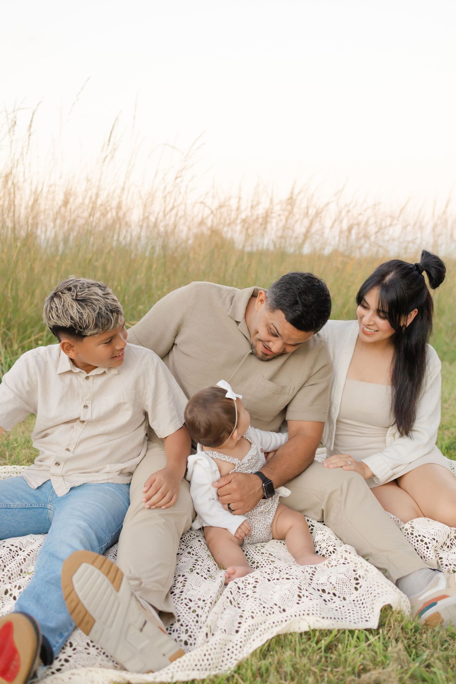 Family of four sitting on a blanket in a grassy field, parents and older child smiling at adorable baby girl.