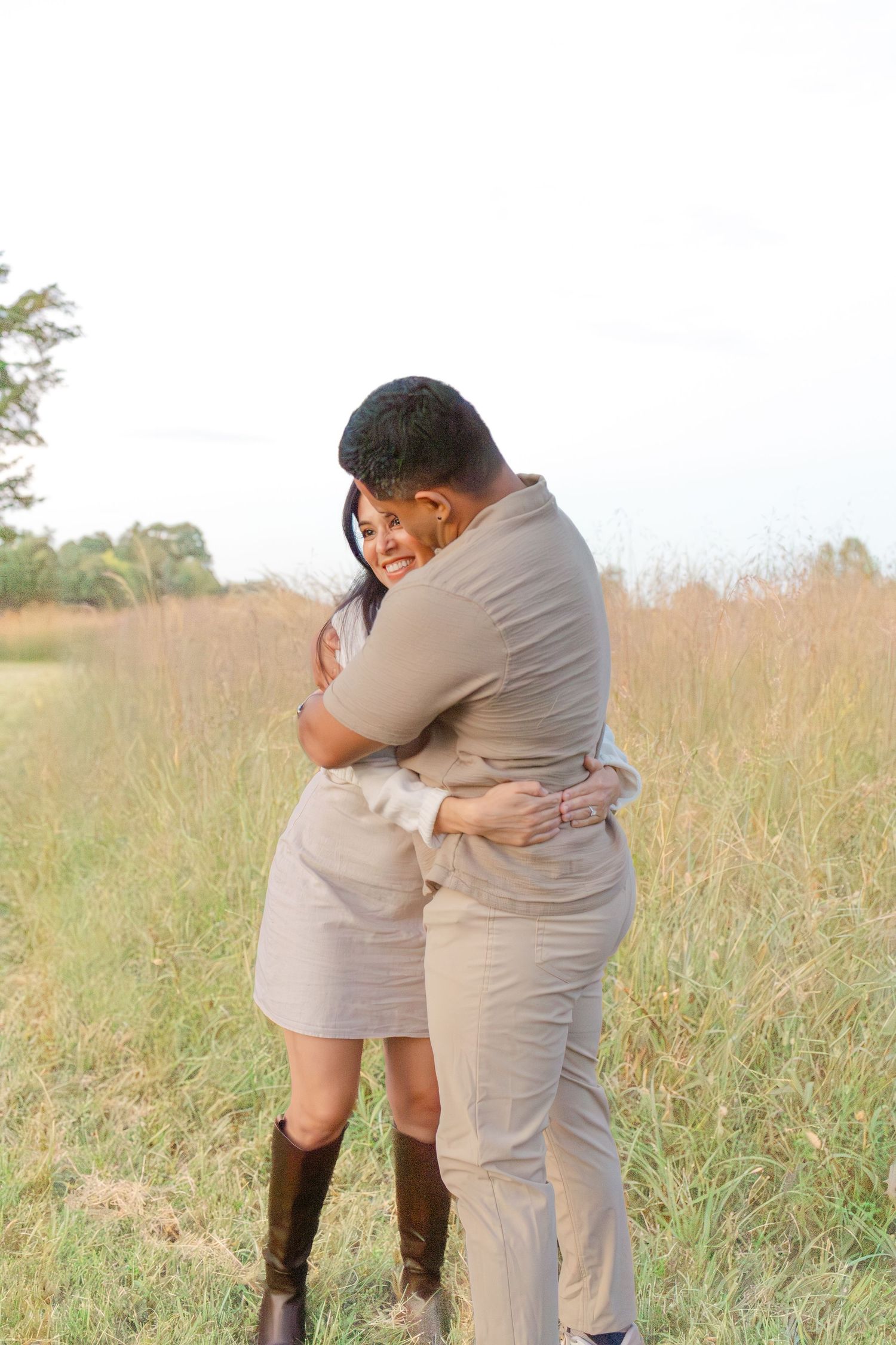Couple embracing in a golden grass field, woman in beige dress and brown cowboy boots, man in khaki outfit.